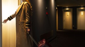 A person in a coat pulling a suitcase, standing in a hallway with doors and a carpeted floor.