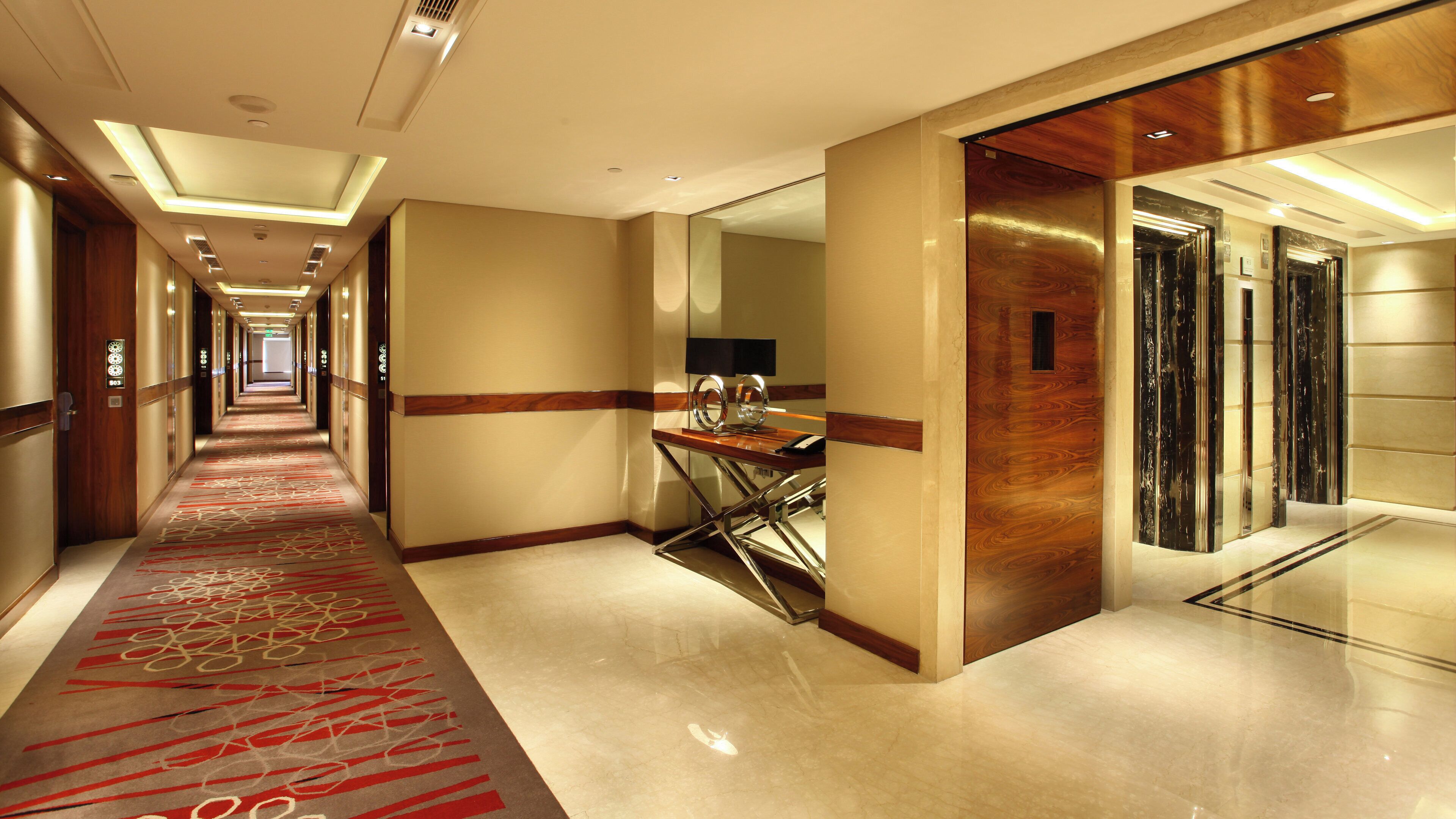 A hotel corridor with a red carpet, a desk with a lamp, and an elevator.