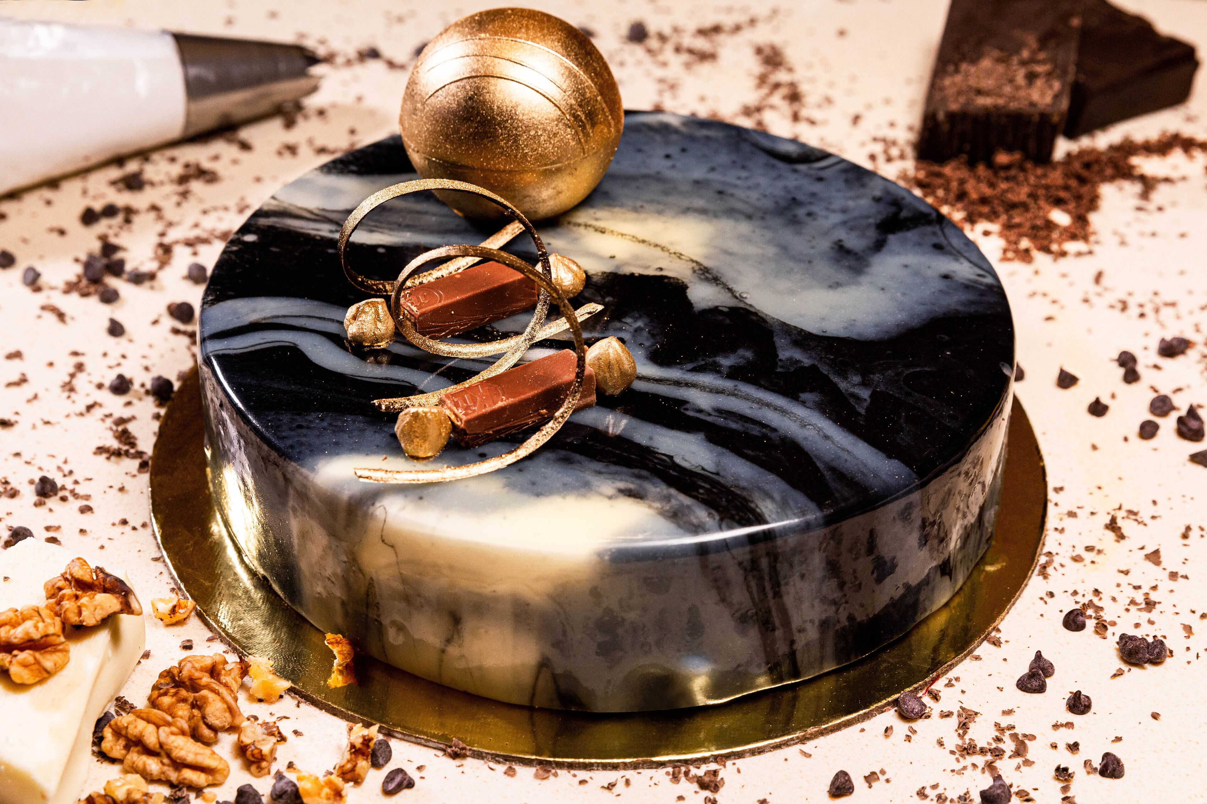 A round cake with a glossy black and white marble finish, topped with a golden ball and chocolate decorations.