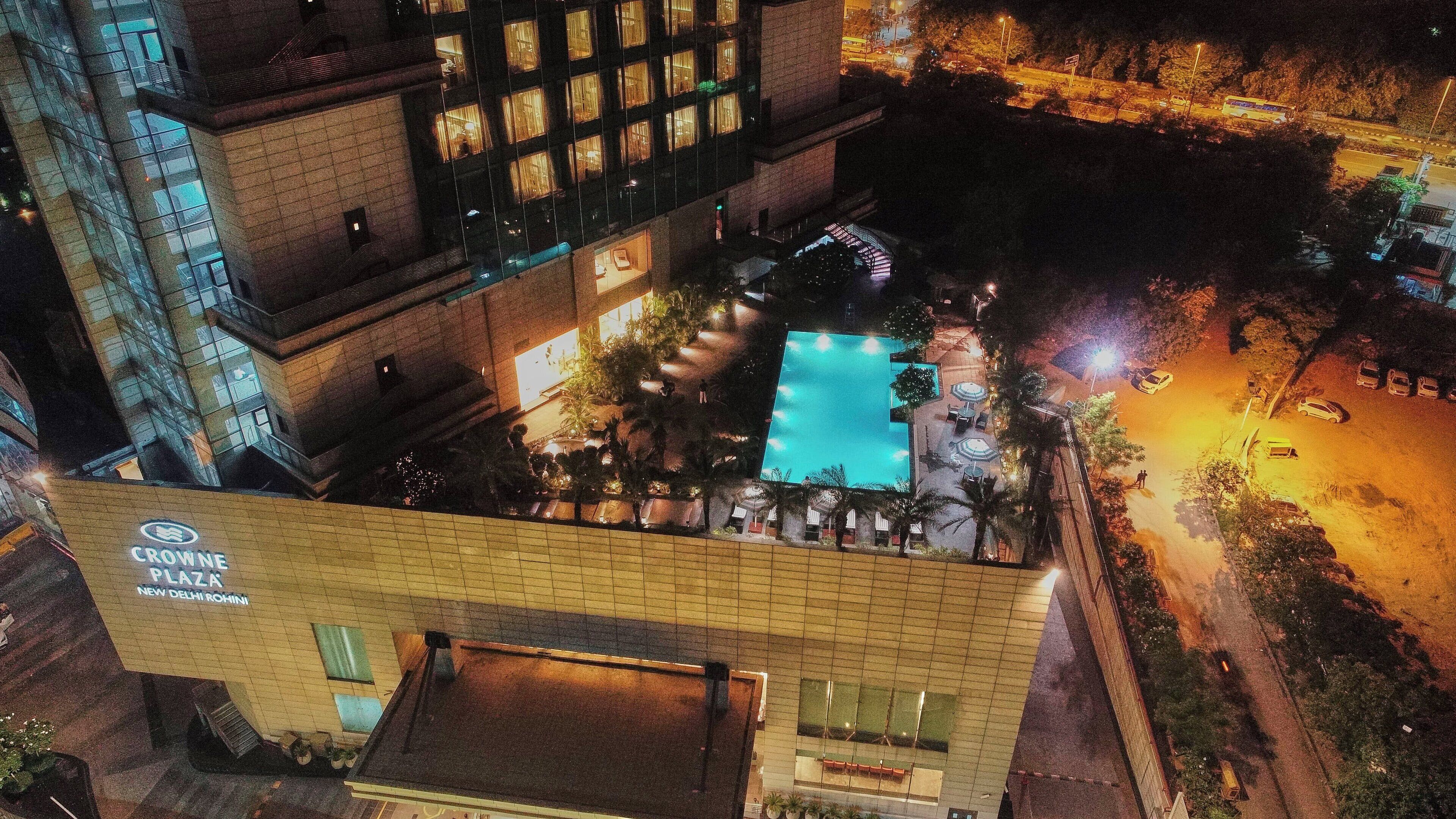 Un hotel amb una piscina al terrat, un cartell que diu 'Crowne Plaza New Delhi Rohini', i una vista de la zona circumstant a la nit.