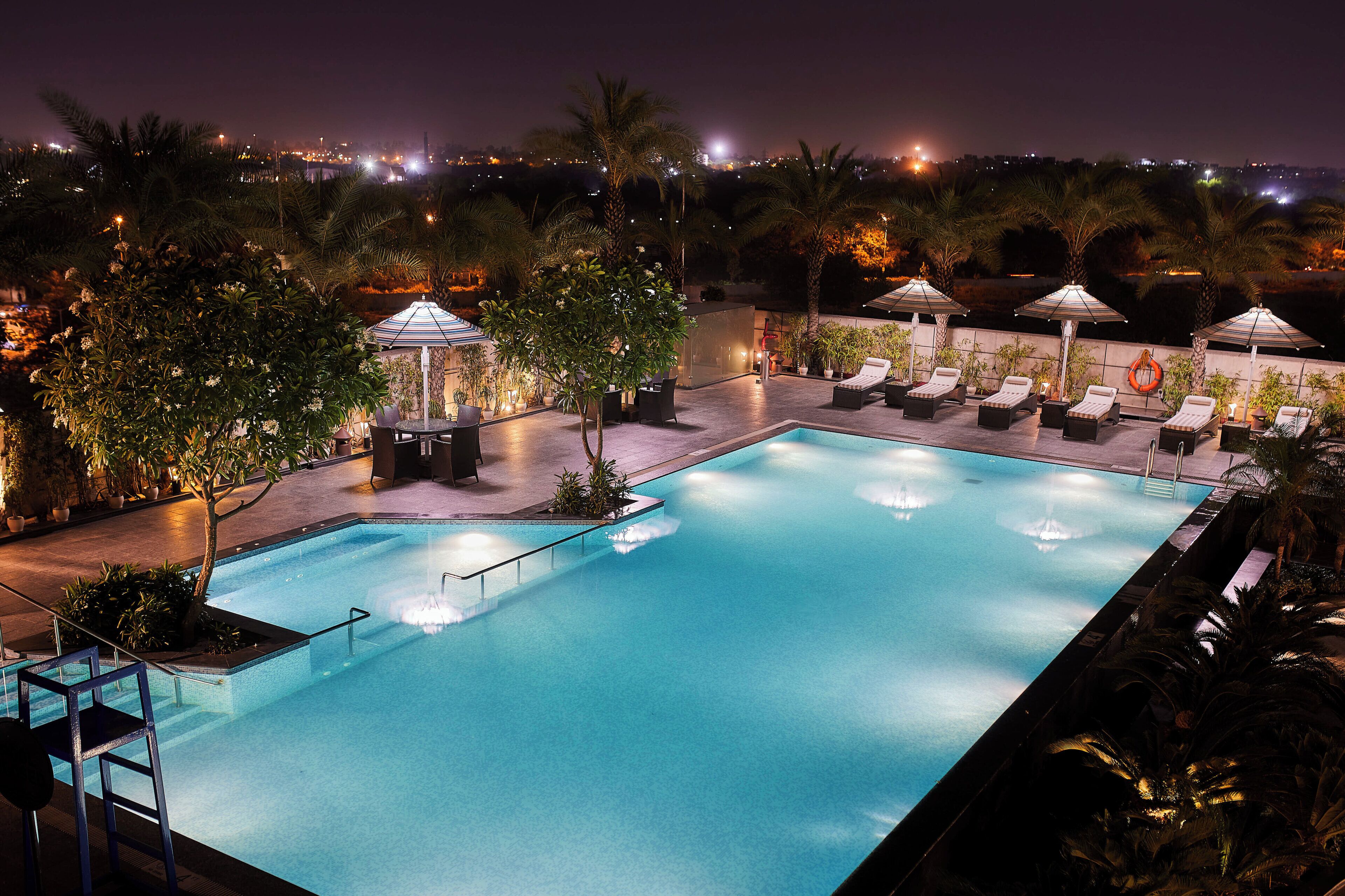 A nighttime swimming pool with lounge chairs and umbrellas, surrounded by palm trees and a cityscape in the background.