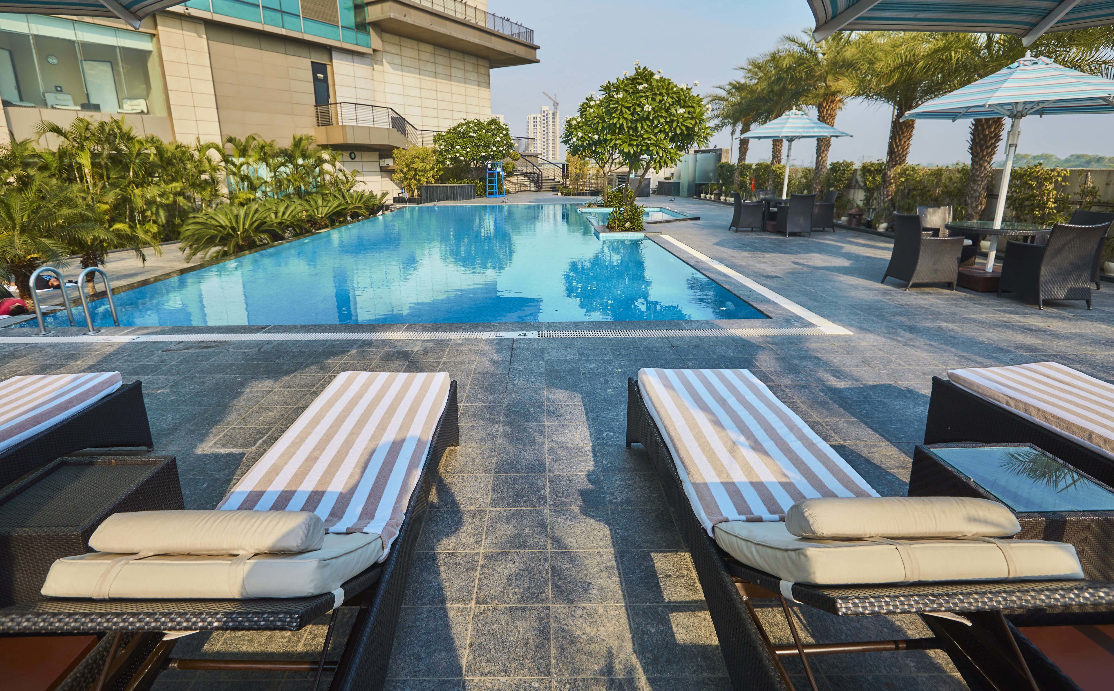 Área de piscina com espreguiçadeiras e guarda-sóis, cercada por vegetação e um edifício ao fundo.