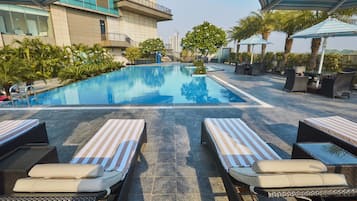 Área de piscina com espreguiçadeiras e guarda-sóis, cercada por vegetação e um edifício ao fundo.