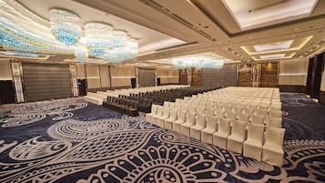Un amplio salón de conferencias con filas de sillas blancas, una alfombra estampada y un techo con candelabros.