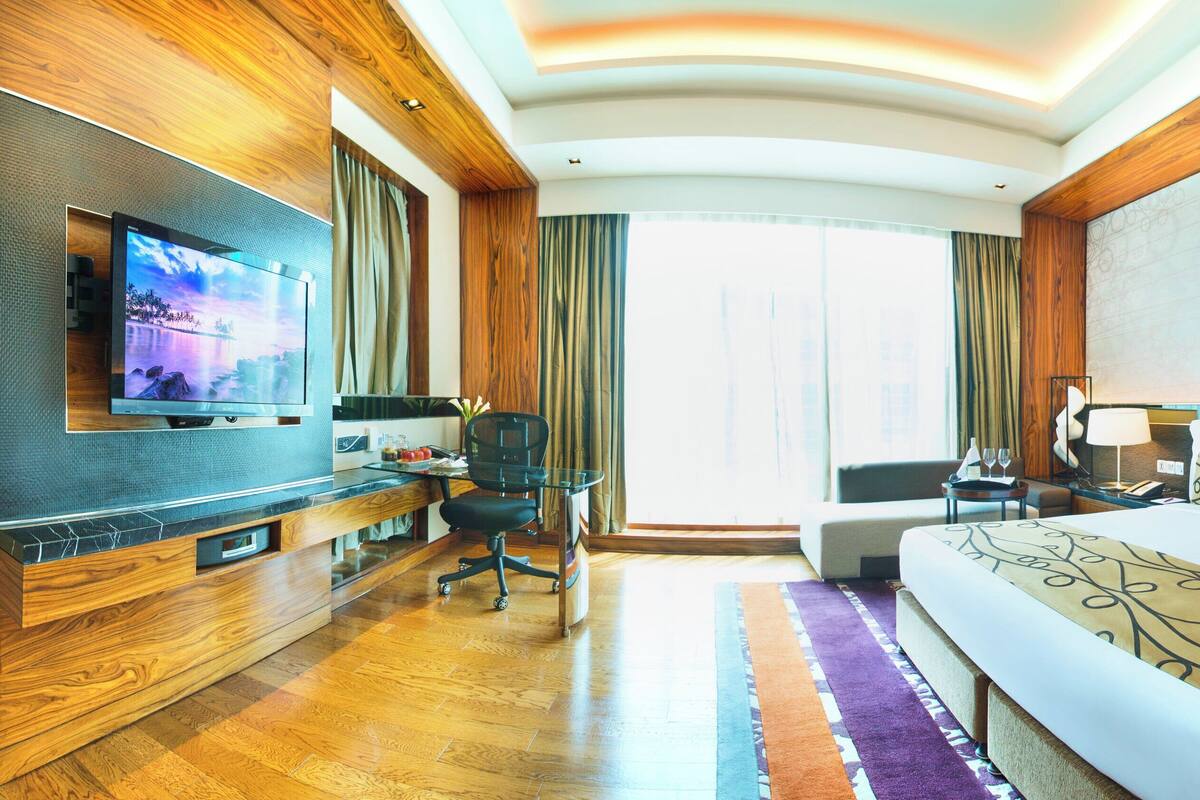 Habitación de hotel con una cama grande, un escritorio con computadora, un televisor y una ventana con cortinas.