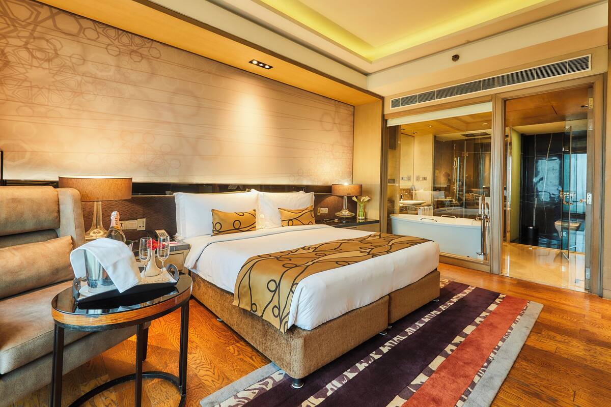 Habitación de hotel con una cama grande, zona de estar con sofá y mesa, baño con bañera y ducha, y puerta de cristal que da a un balcón.