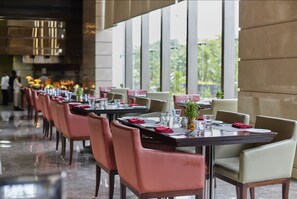A restaurant with long tables, red napkins, and a view of greenery through large windows.