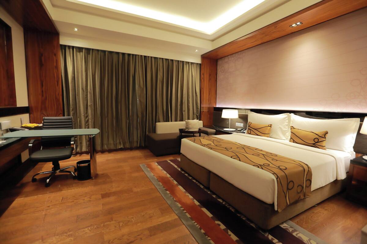 Habitación de hotel con una cama grande, un escritorio con silla, una mesita y una mesita de noche con lámpara.