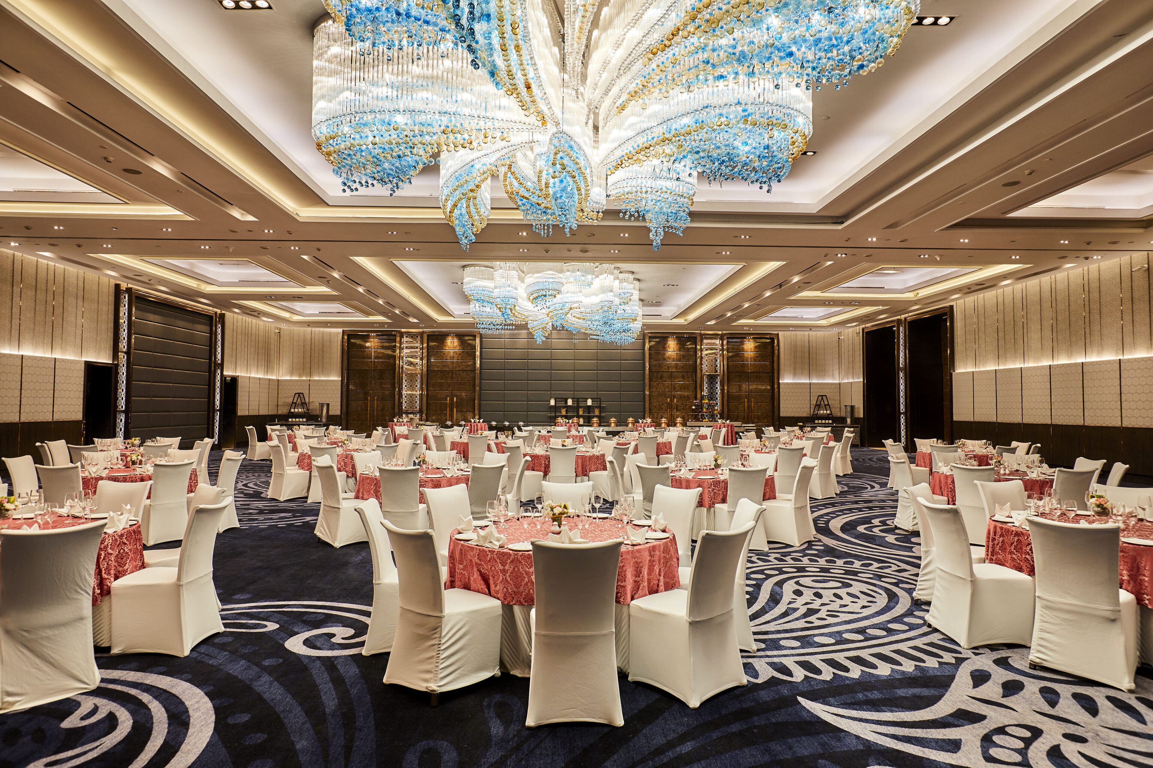 Un amplio salón de banquetes con mesas redondas, sillas blancas y una lámpara de araña imponente.