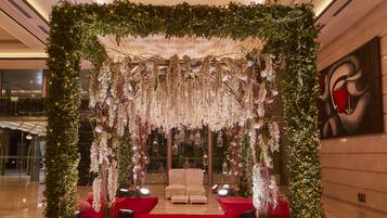 A luxurious indoor setting with a red carpet leading to a white sofa and chairs, surrounded by greenery and hanging floral decorations.