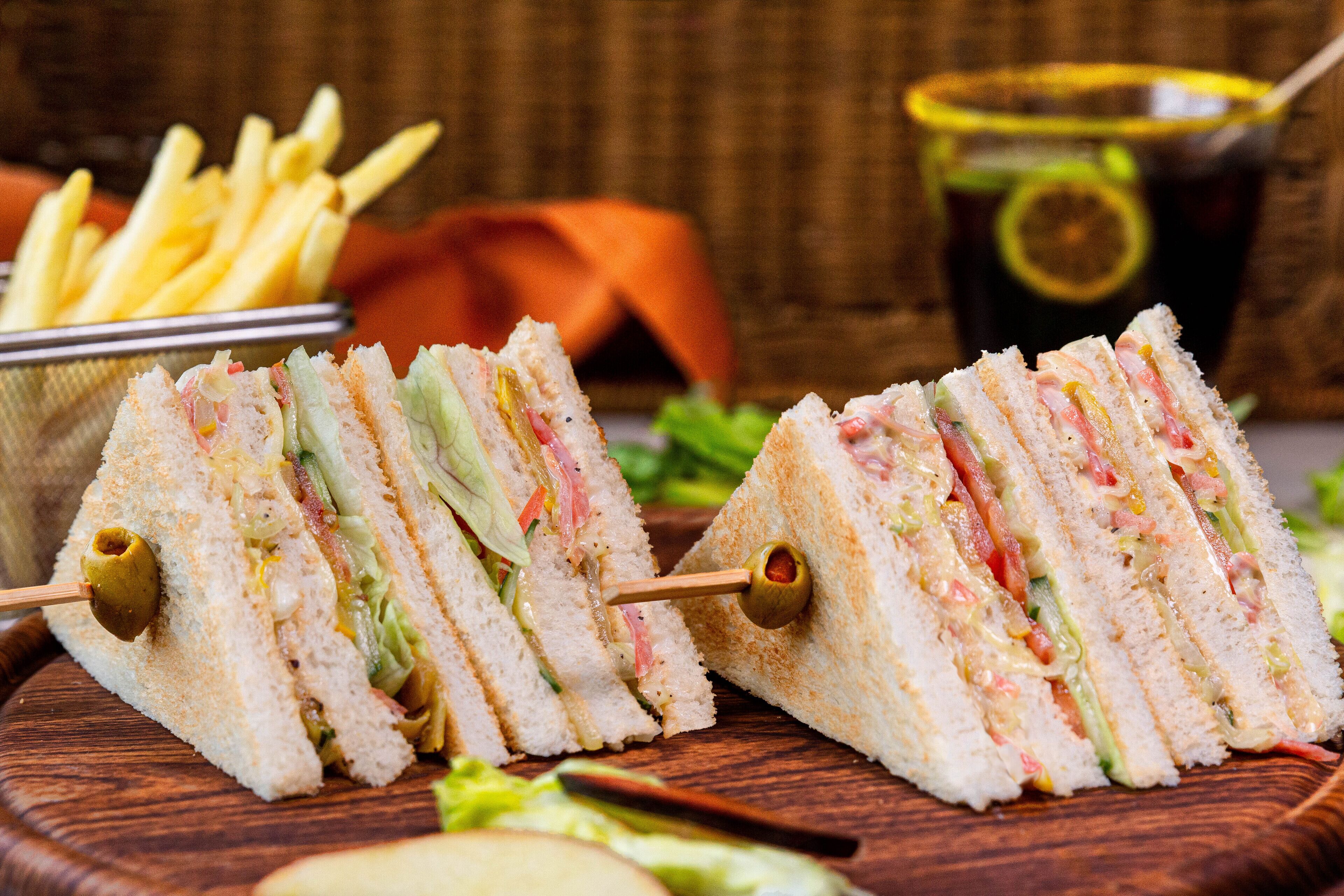 A sandwich cut into triangles with an olive on a stick, served with fries and a drink in the background.