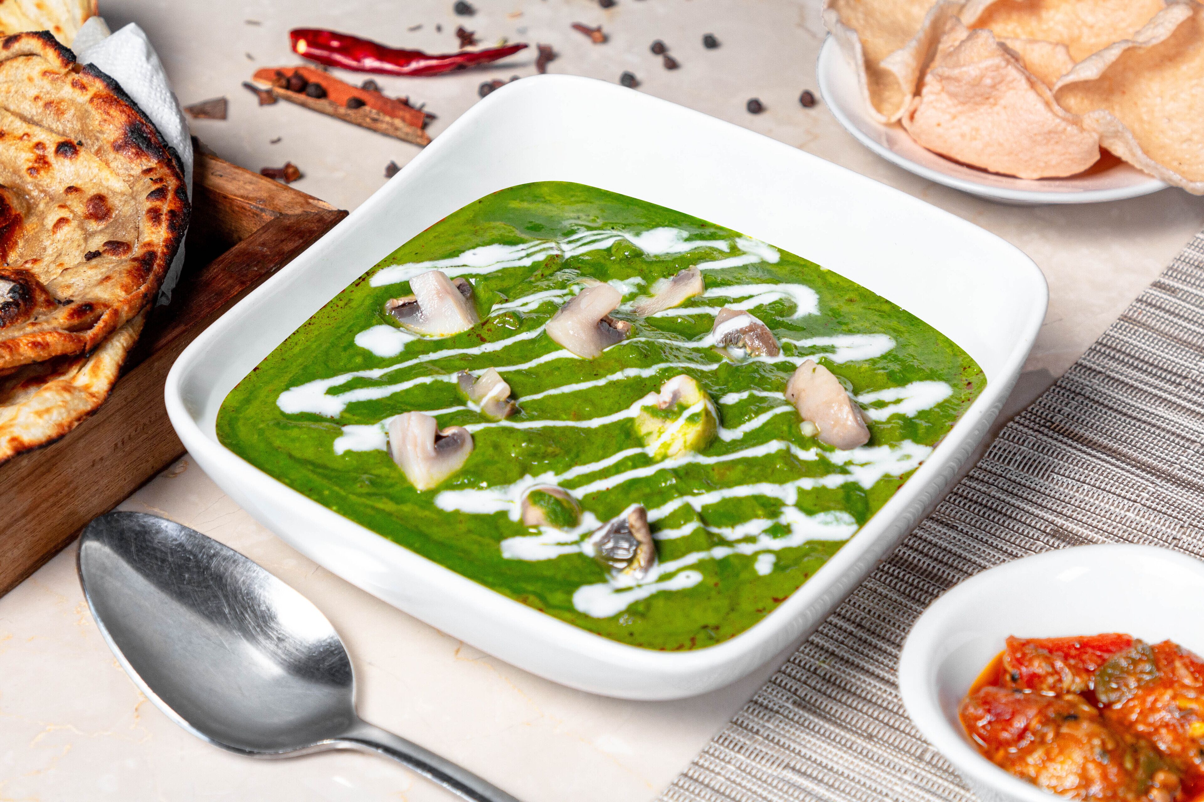 A bowl of green soup with white sauce and mushrooms, accompanied by flatbread and a small bowl of red sauce.