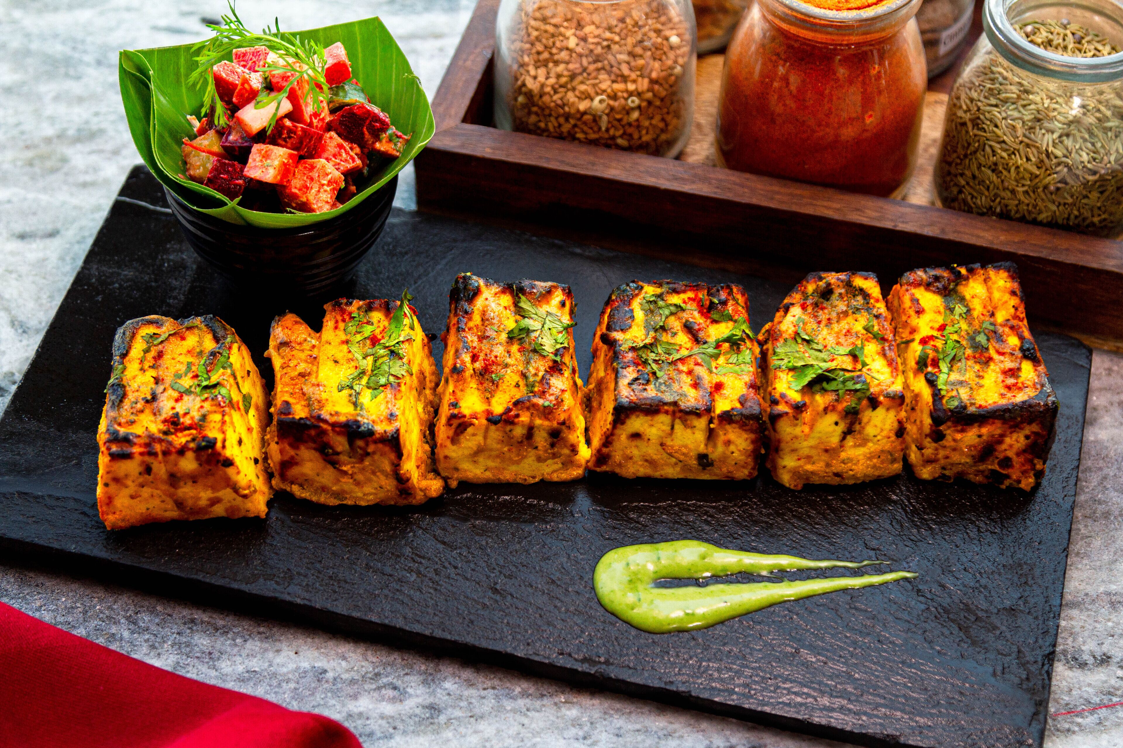 A black slate serving board with grilled paneer cubes garnished with herbs, a bowl of mixed fruit salad, and a small jar of green chutney.