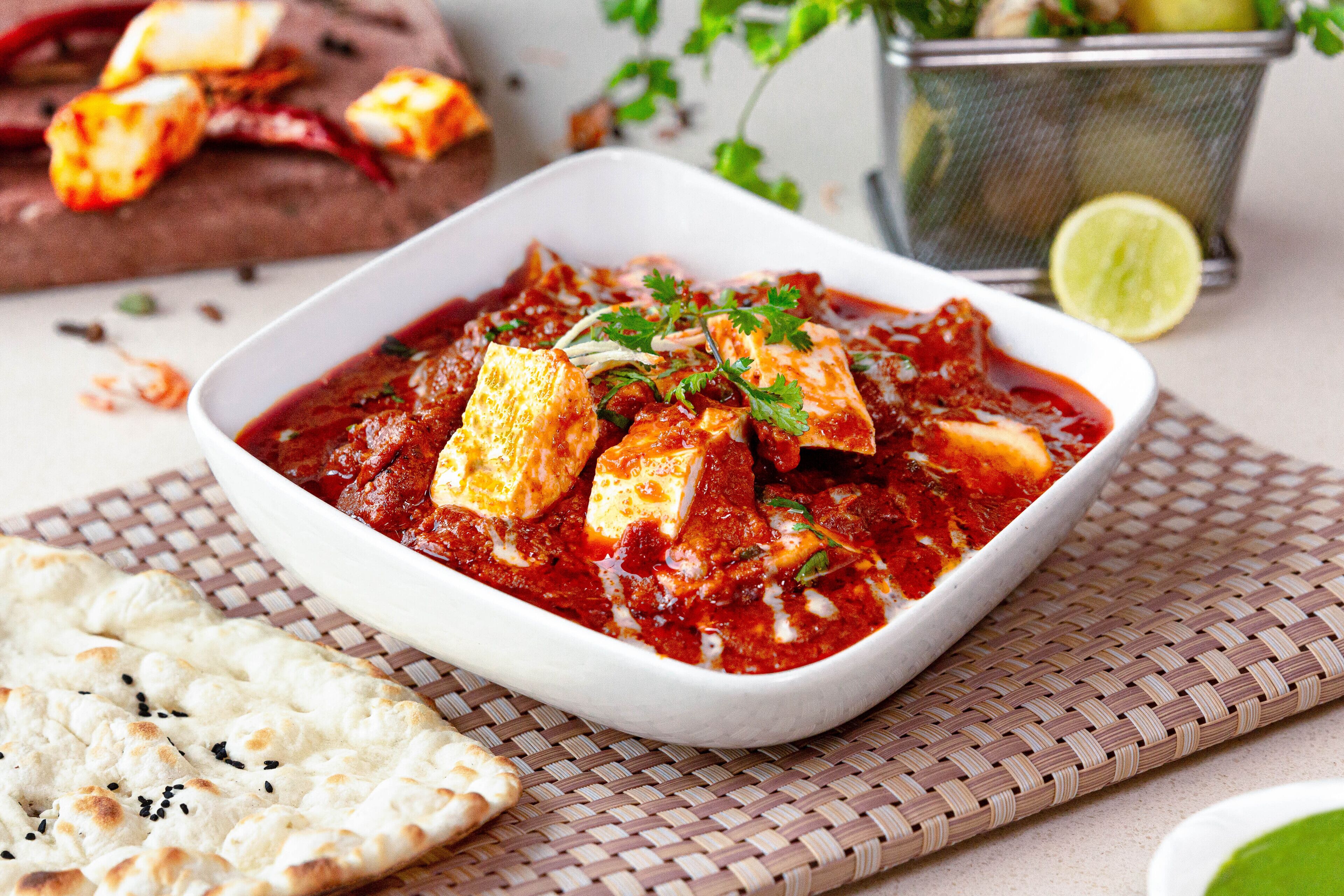 A white bowl of paneer curry with paneer cubes, garnished with cilantro, served with naan bread on a woven mat.