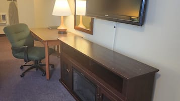 A hotel room with a flat-screen TV mounted on the wall, a wooden desk with a lamp, a green office chair, and a fireplace.