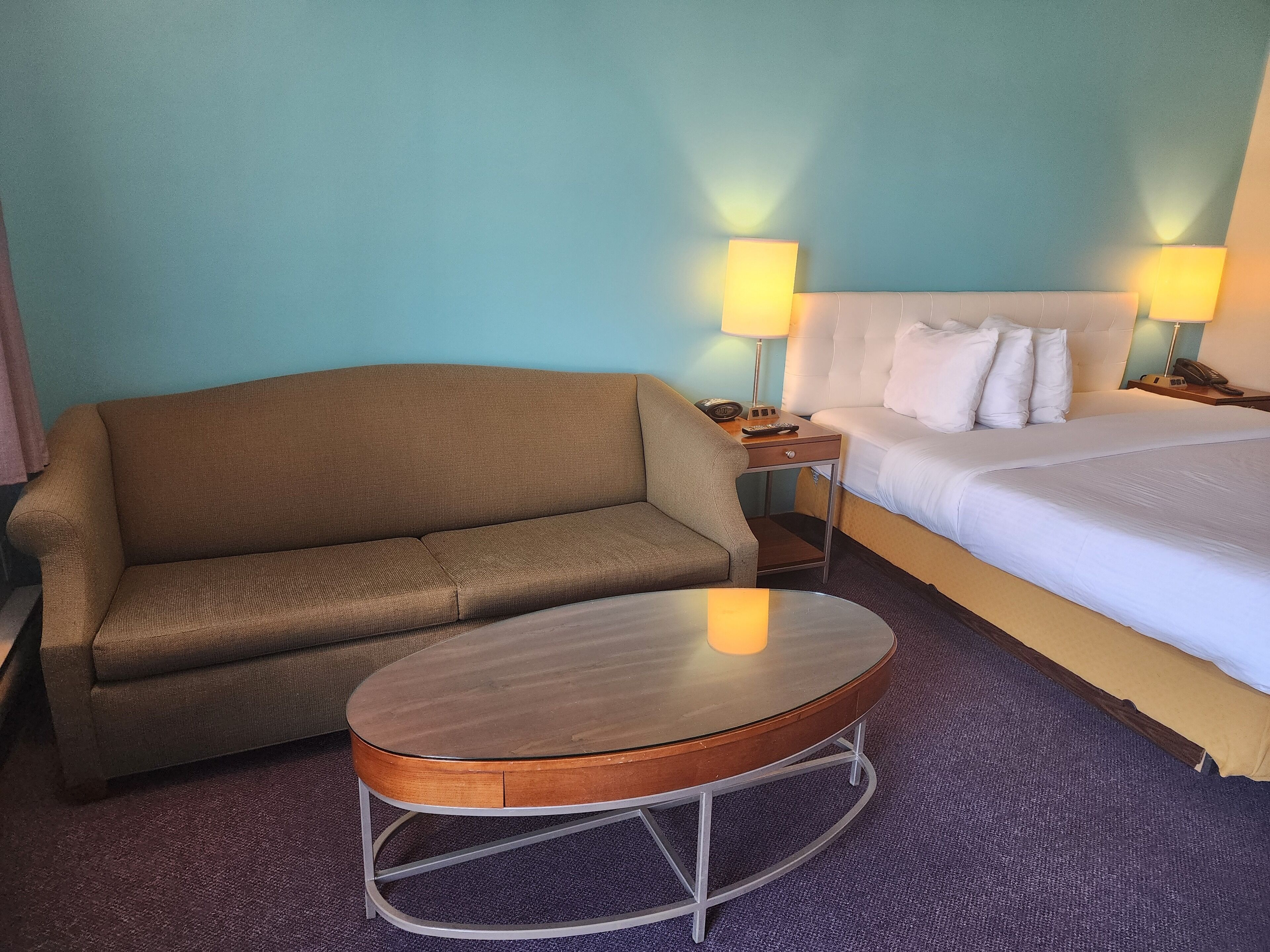 A hotel room with a brown sofa, a round wooden table, a bed with white bedding, and two bedside lamps.