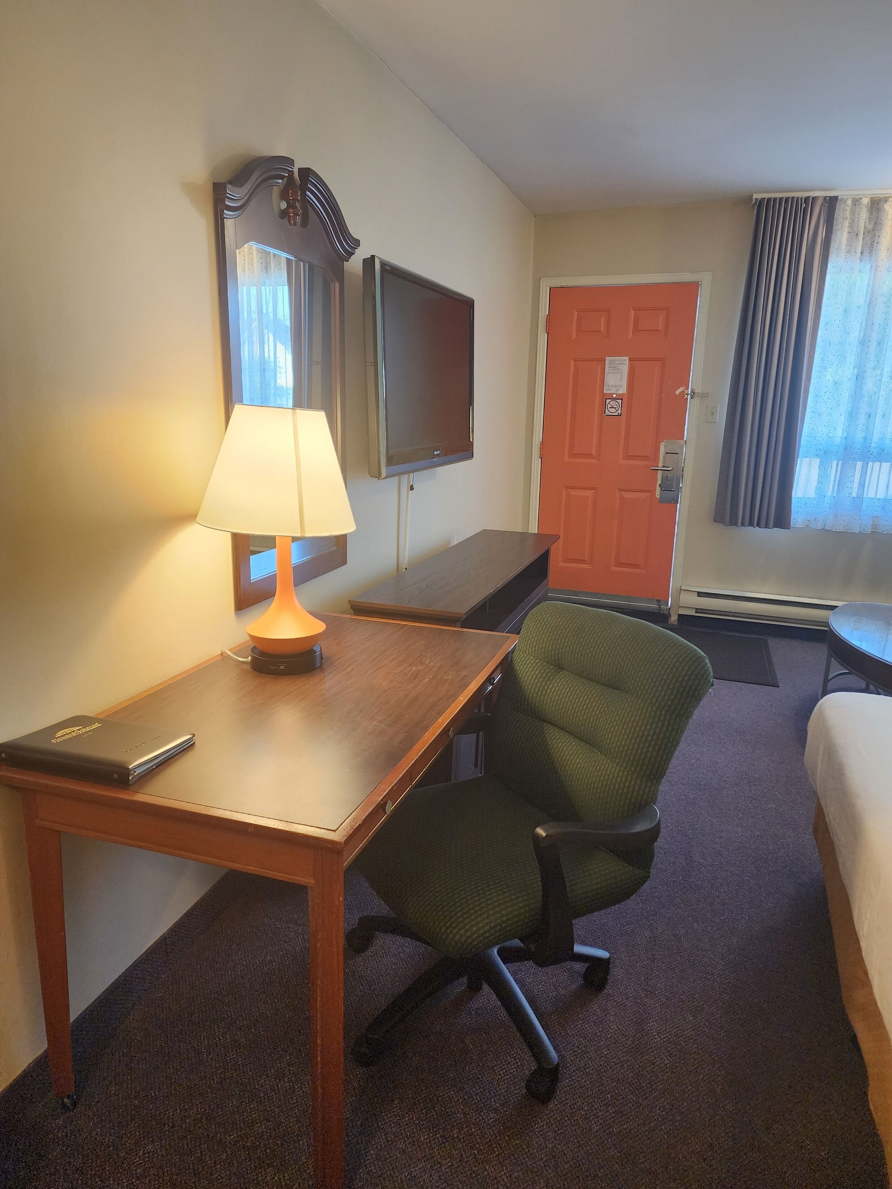 A hotel room with a wooden desk, a green office chair, a TV mounted on the wall, and a bed with a white comforter.