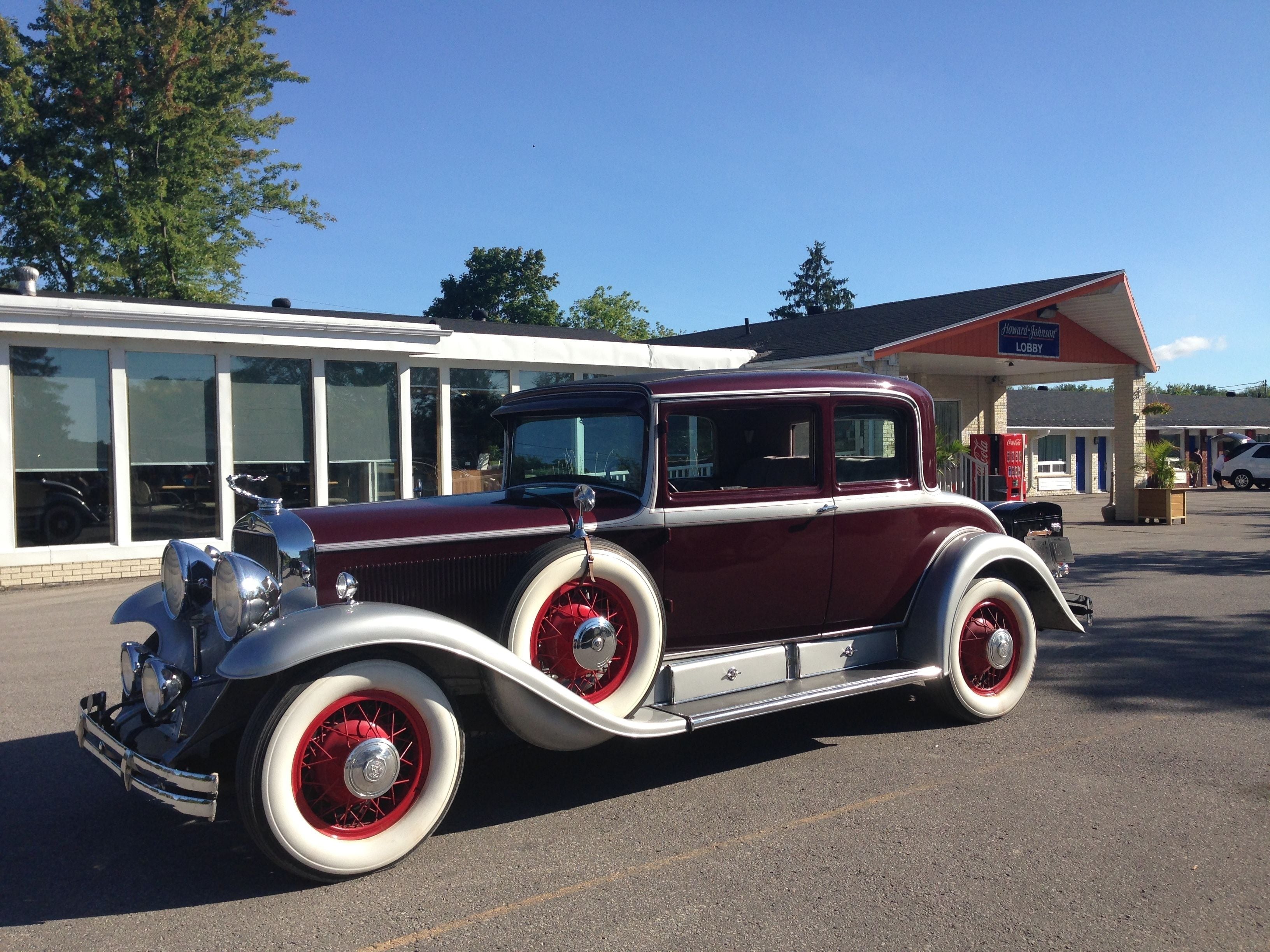 A vintage car parked in front of a building with large windows and a Coca-Cola vending machine.