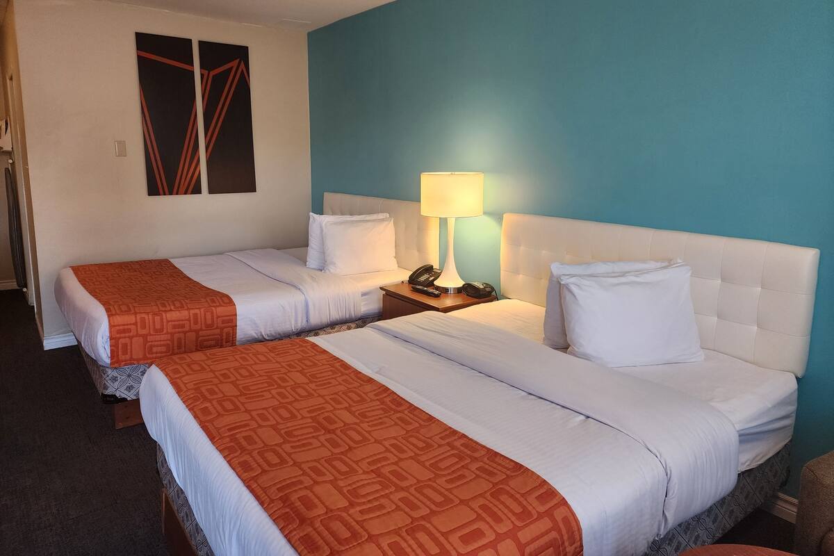 Habitación de hotel con dos camas, cada una con sábanas blancas y colchas estampadas anaranjadas. Entre las camas, un mesita de noche con lámpara y teléfono. La habitación tiene una pared azul decorativa y un cuadro sobre las camas.