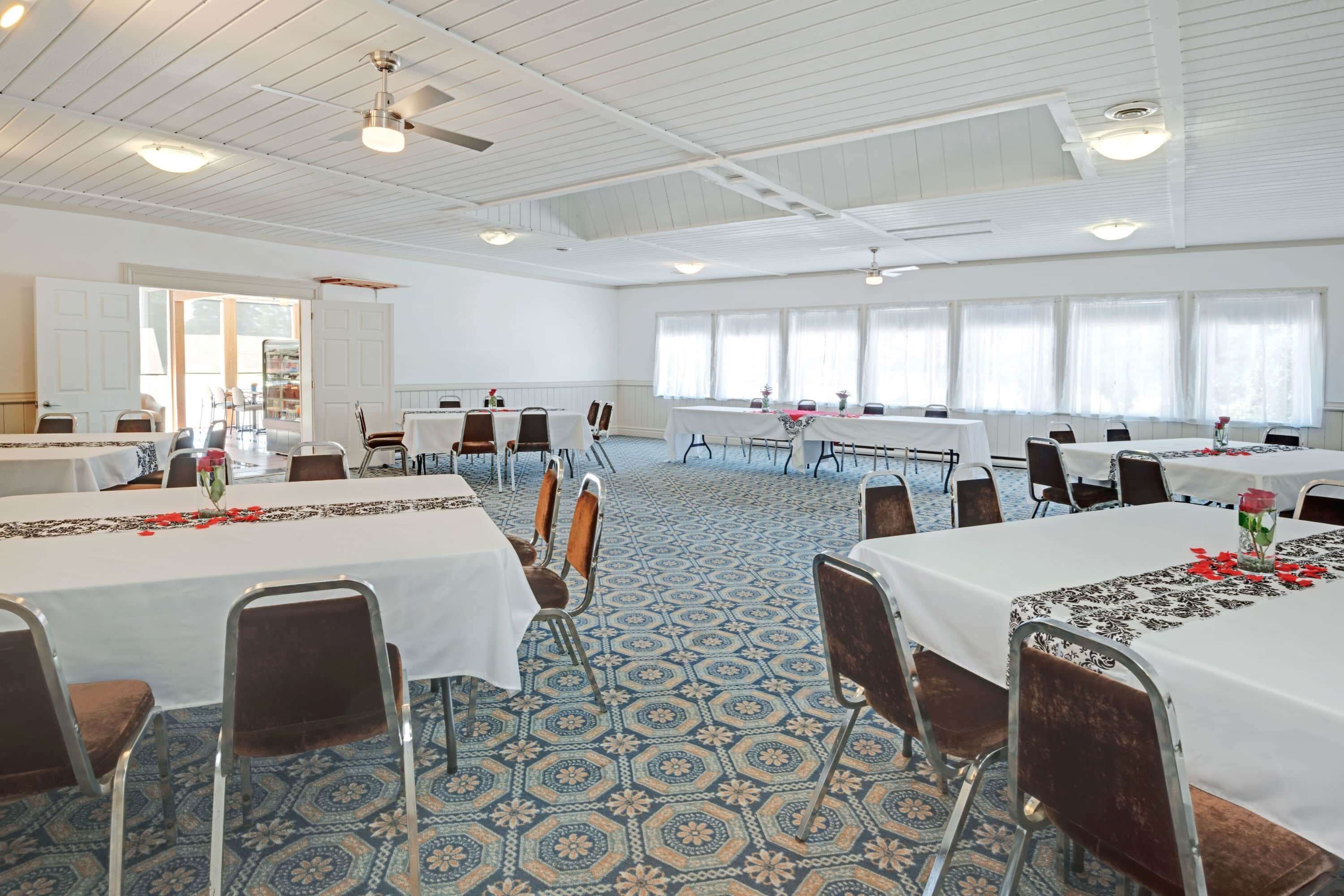 A spacious banquet hall with multiple tables and chairs, white ceiling with exposed beams, and patterned carpet.