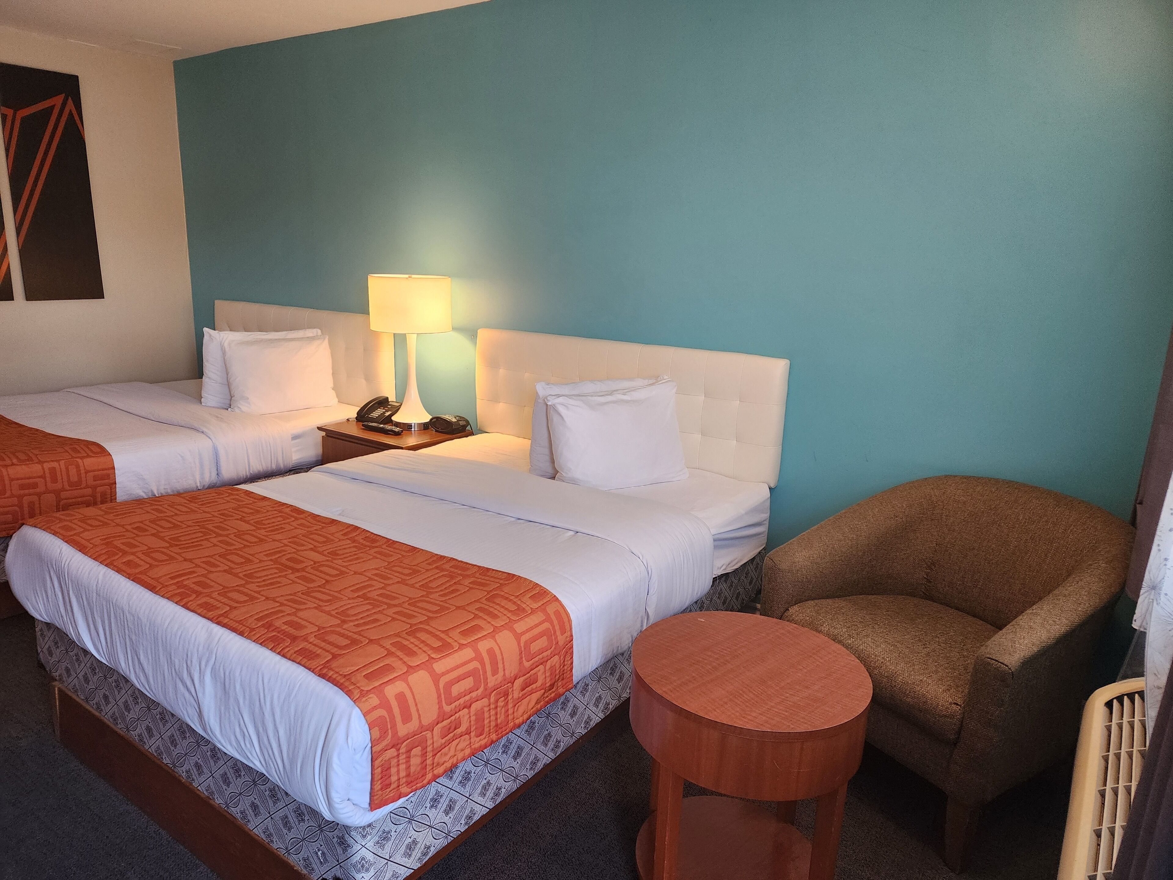 A hotel room with two beds, a round wooden side table, a brown armchair, and a lamp on a nightstand.