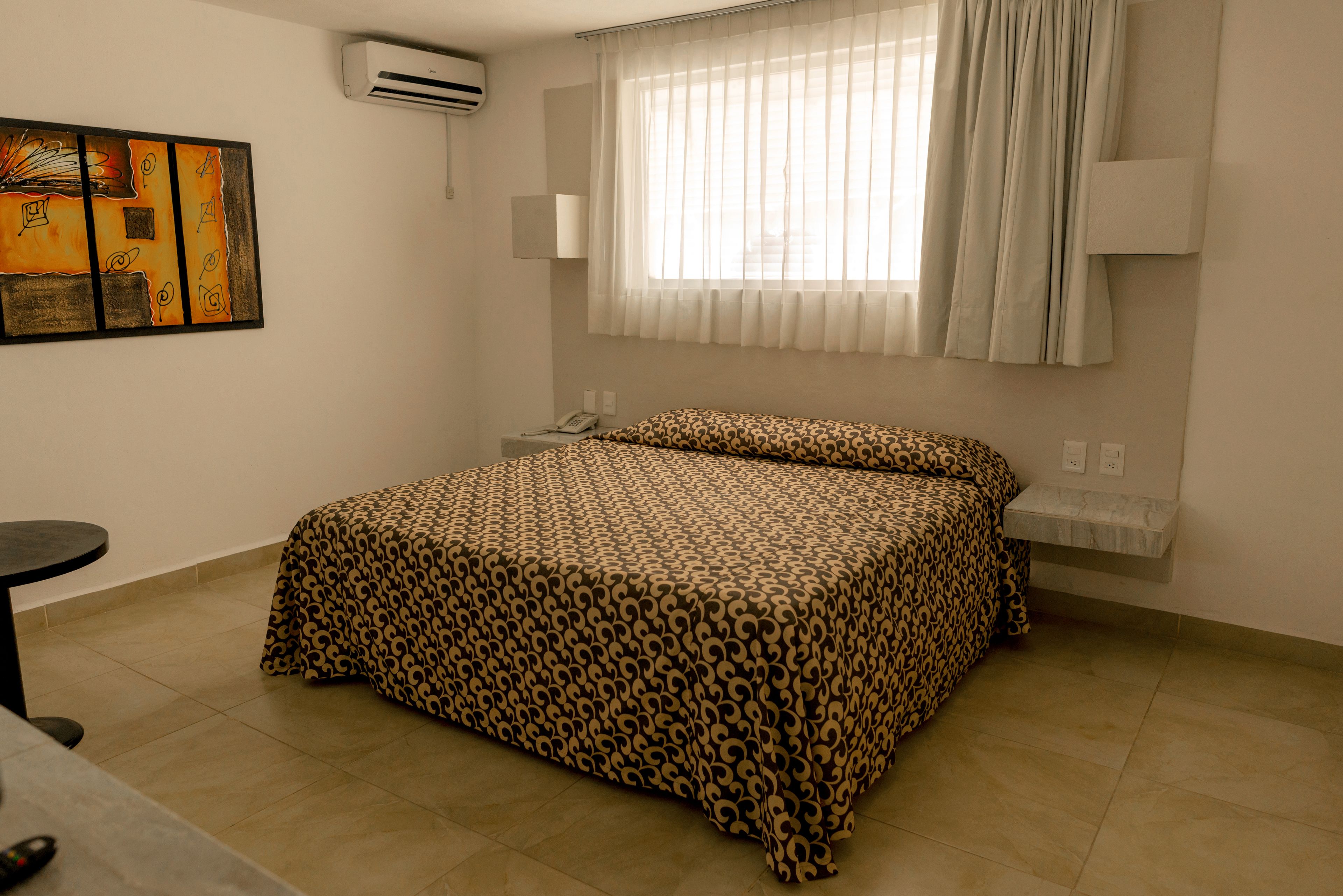 Una habitación de hotel con cama, mesita de noche, lámpara, ventana con cortinas y un equipo de aire acondicionado.