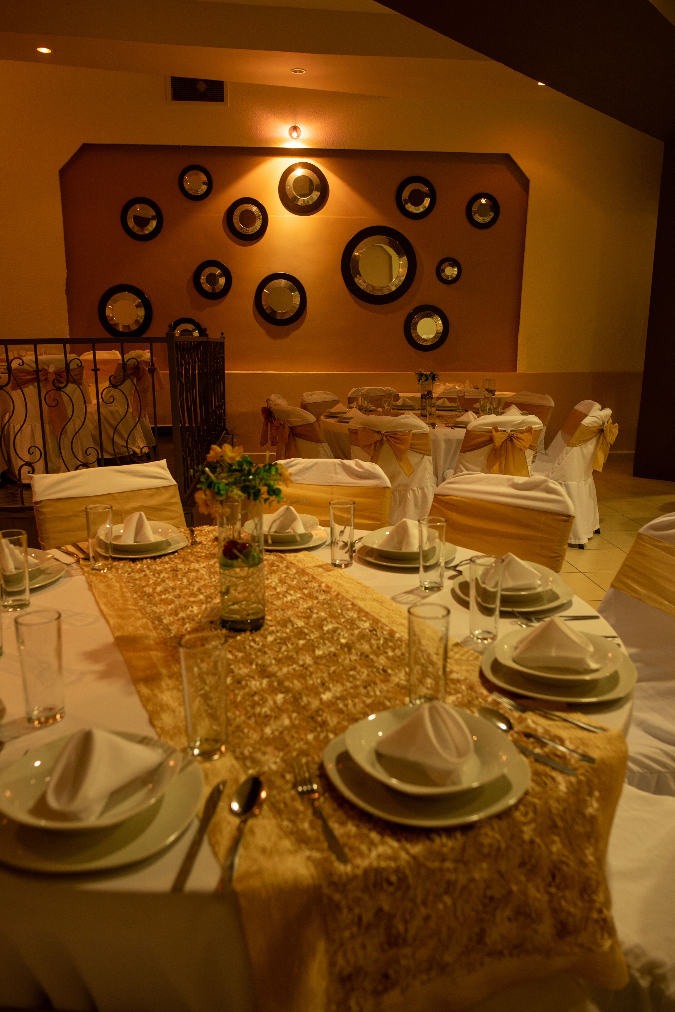 Een balzaal met ronde tafels, gedekt voor een formeel evenement, met gouden tafelkleden, witte serviesgoed en een decoratieve wand met ronde spiegels.