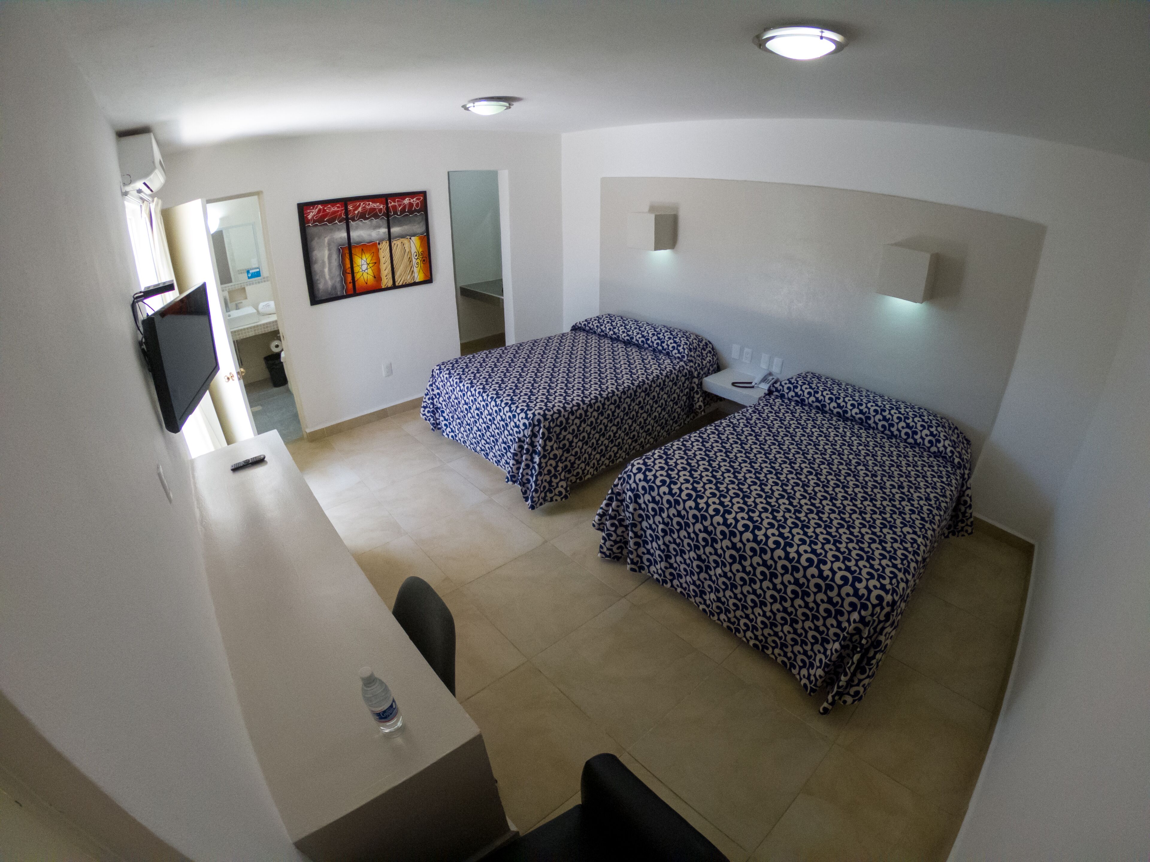 A hotel room with two beds, a desk, a chair, a TV, and a bathroom visible in the background.