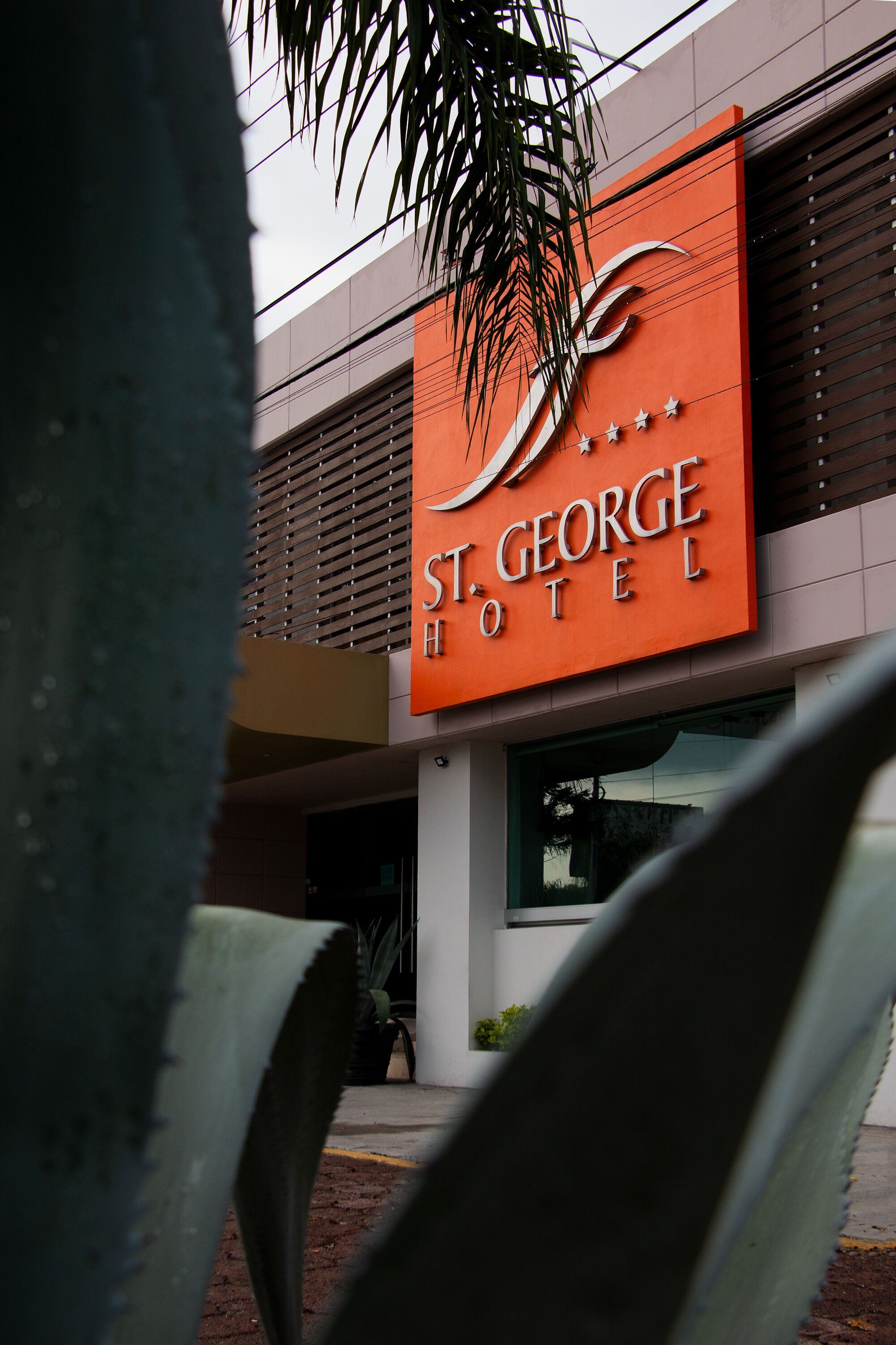Exterior del Hotel St. George, con un llamativo letrero naranja, arquitectura moderna y una palmera en primer plano.