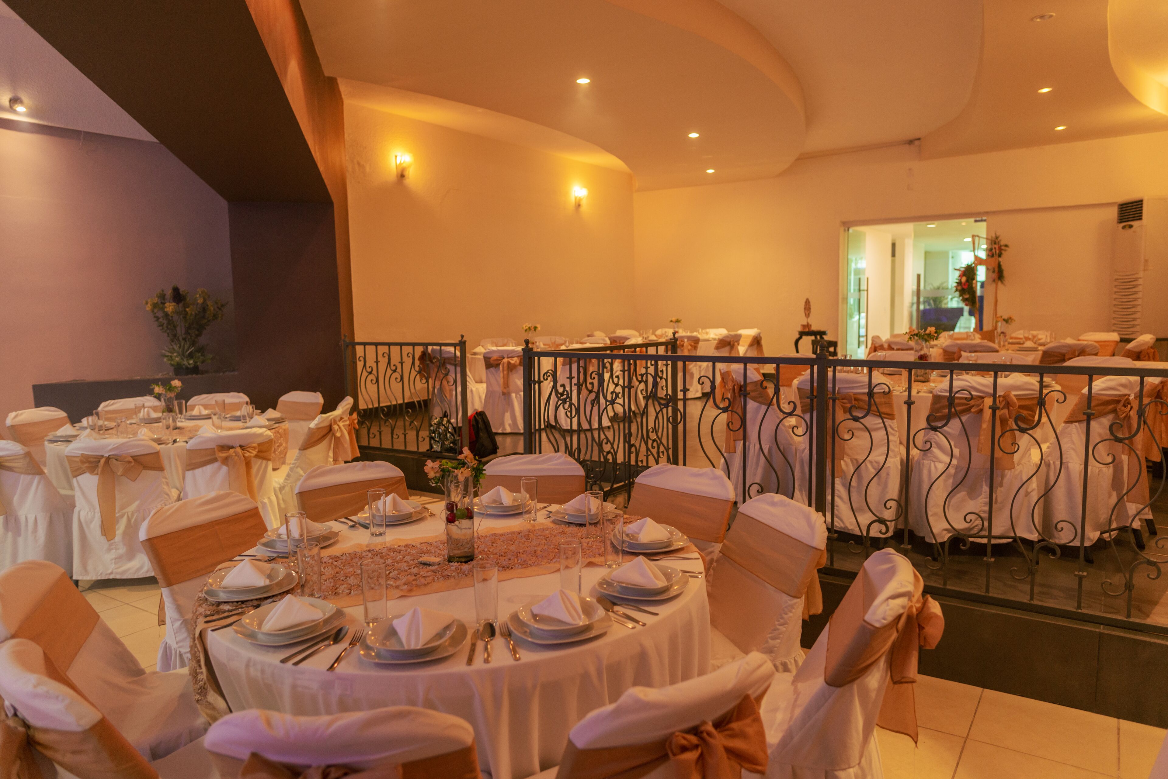 Un salón de banquetes con mesas redondas dispuestas para un evento formal, con manteles blancos, fajas doradas y centros de mesa florales.