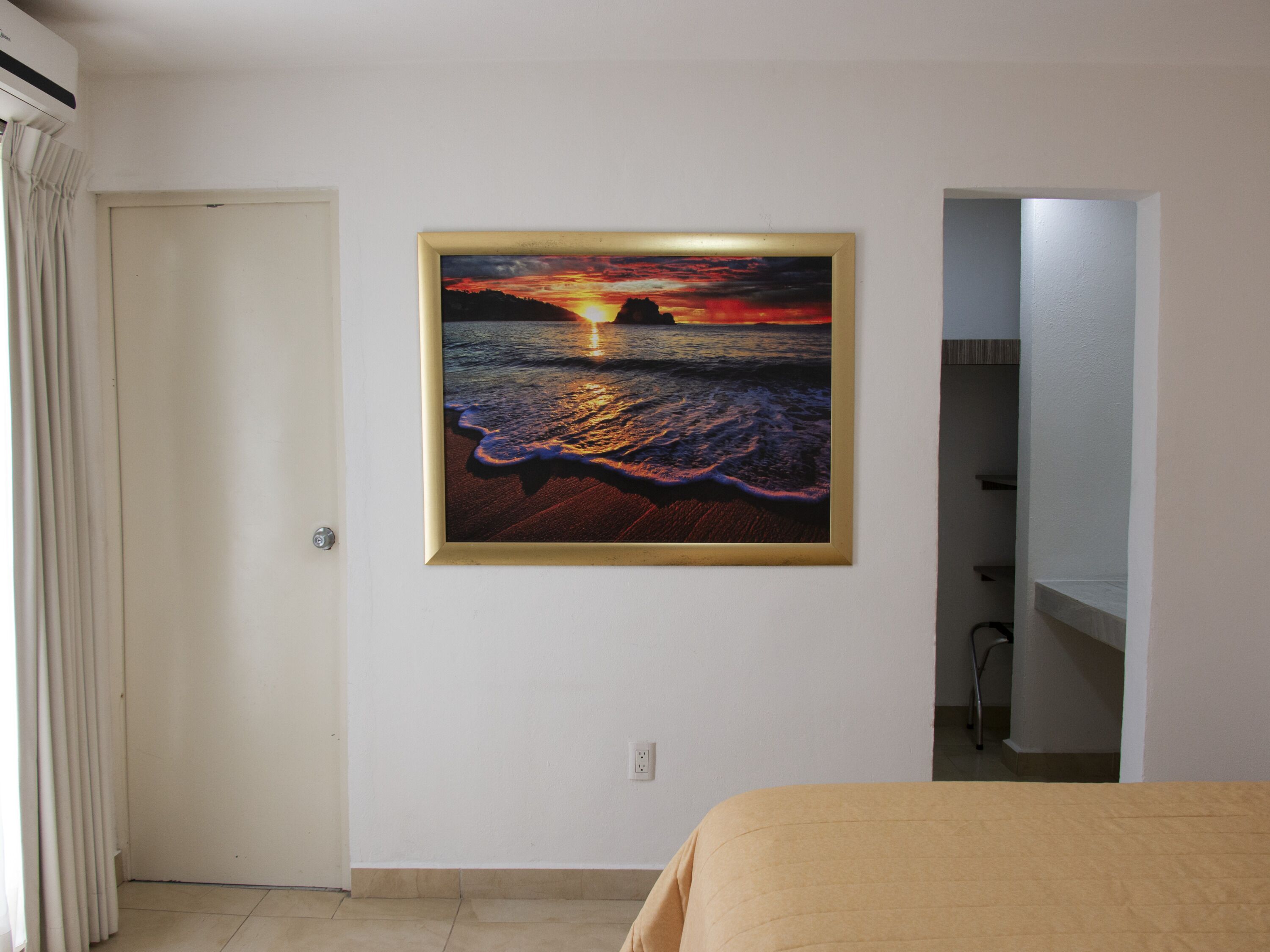 Una habitación con una cama, un cuadro de un atardecer sobre el océano, una puerta y un baño visible a través de un vano abierto.