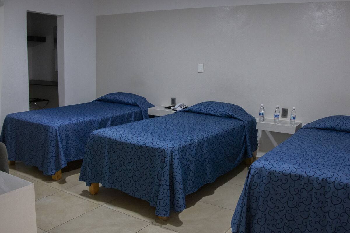 A hotel room with three single beds, each with a blue patterned bedspread and matching pillows. There is a white bedside table with a phone and a water bottle on it. The room has a tiled floor and a white wall.