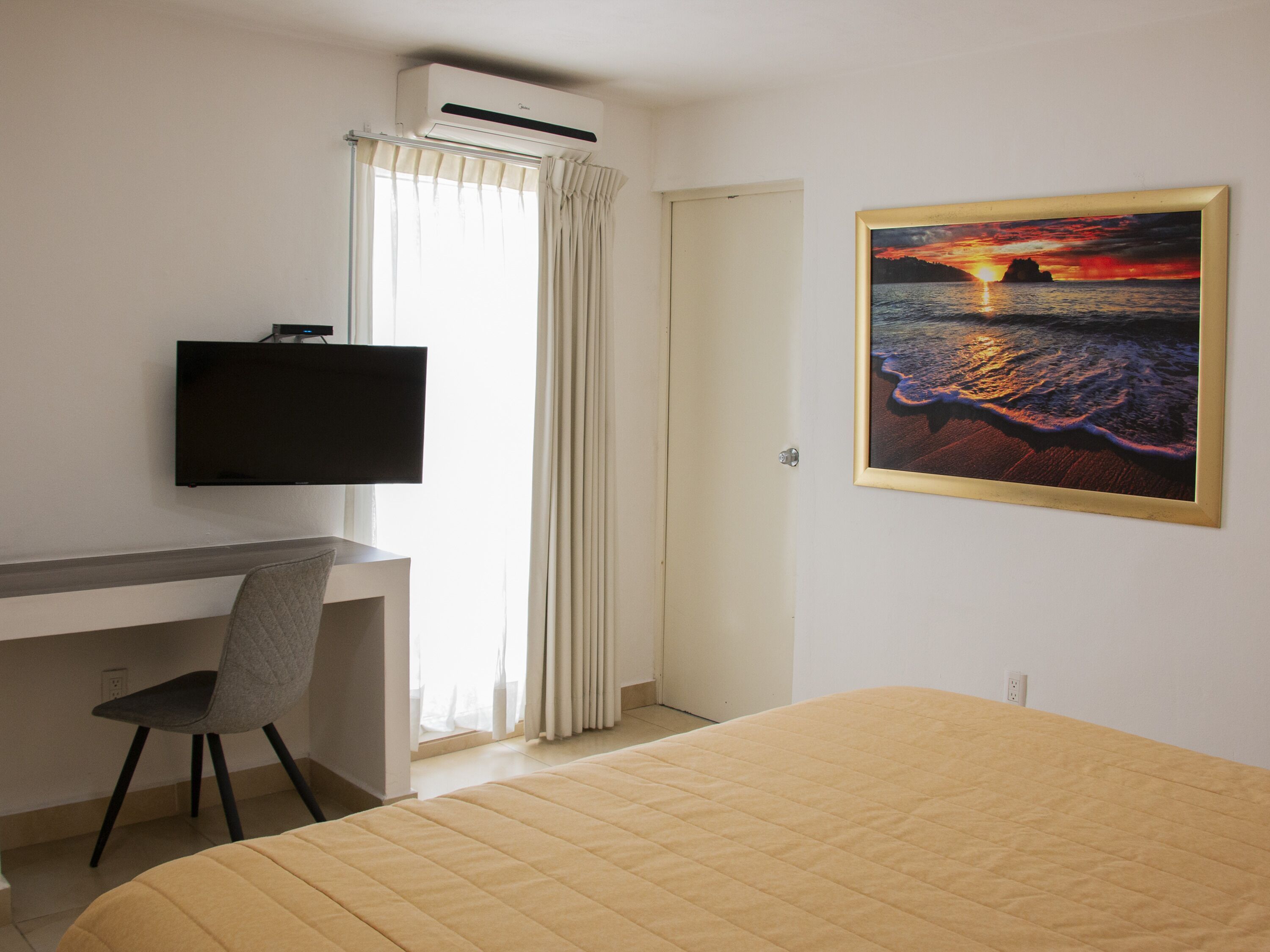 Una habitación de hotel con una cama, un escritorio con una silla, un televisor, un cuadro de un atardecer y un equipo de aire acondicionado.