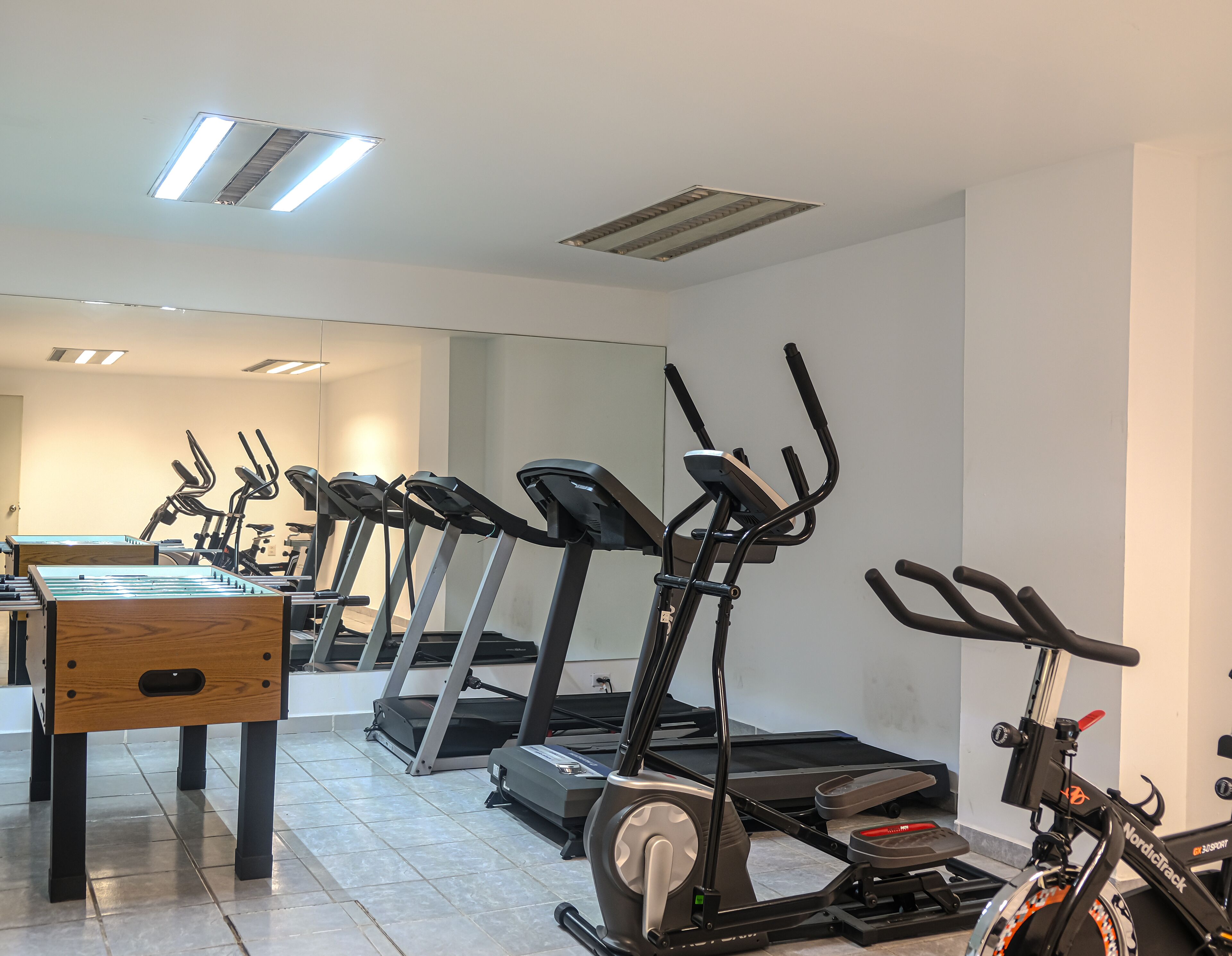 Een sportschool met meerdere loopbanden en hometrainers, een houten tafel en een spiegel aan de muur.