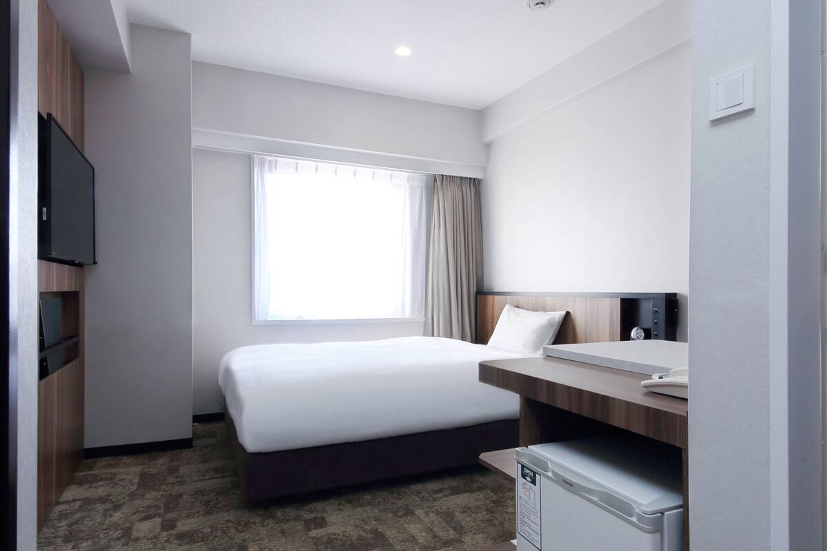 Une chambre d’hôtel équipée d’un lit, d’un bureau, d’un petit réfrigérateur et d’un téléviseur fixé au mur.