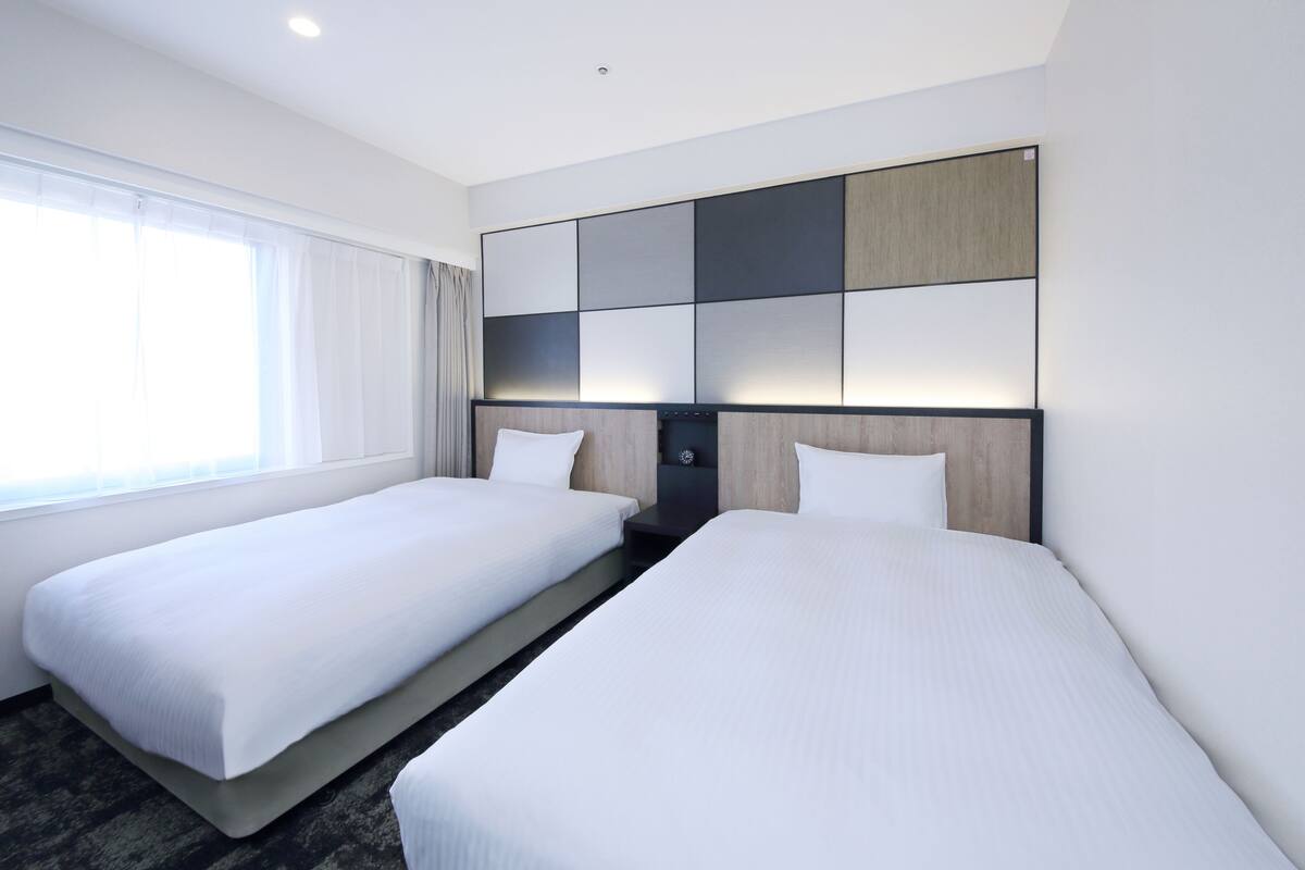 Une chambre d’hôtel avec deux lits simples, une grande fenêtre avec des rideaux et une tête de lit moderne dotée d’une horloge intégrée.