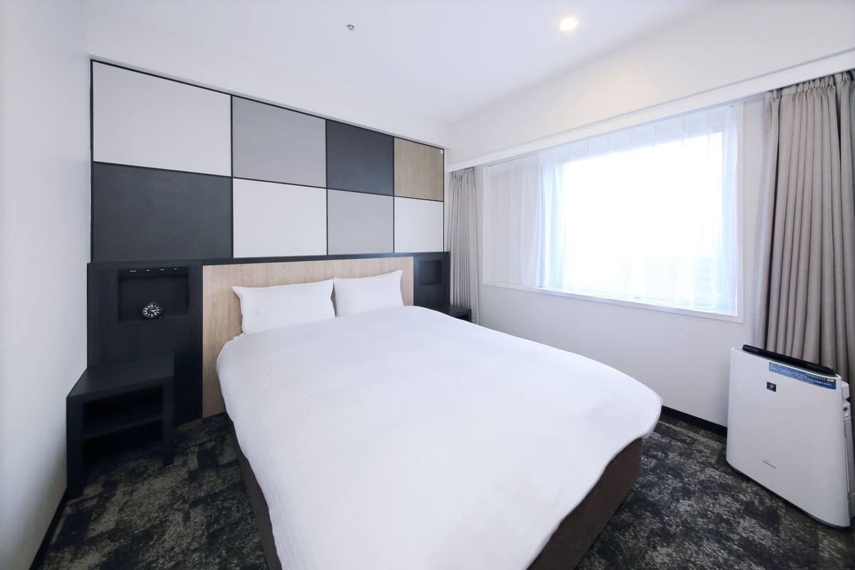 Une chambre d’hôtel avec un grand lit, une tête de lit intégrant un téléviseur, une table de chevet, une fenêtre avec des rideaux et un radiateur.