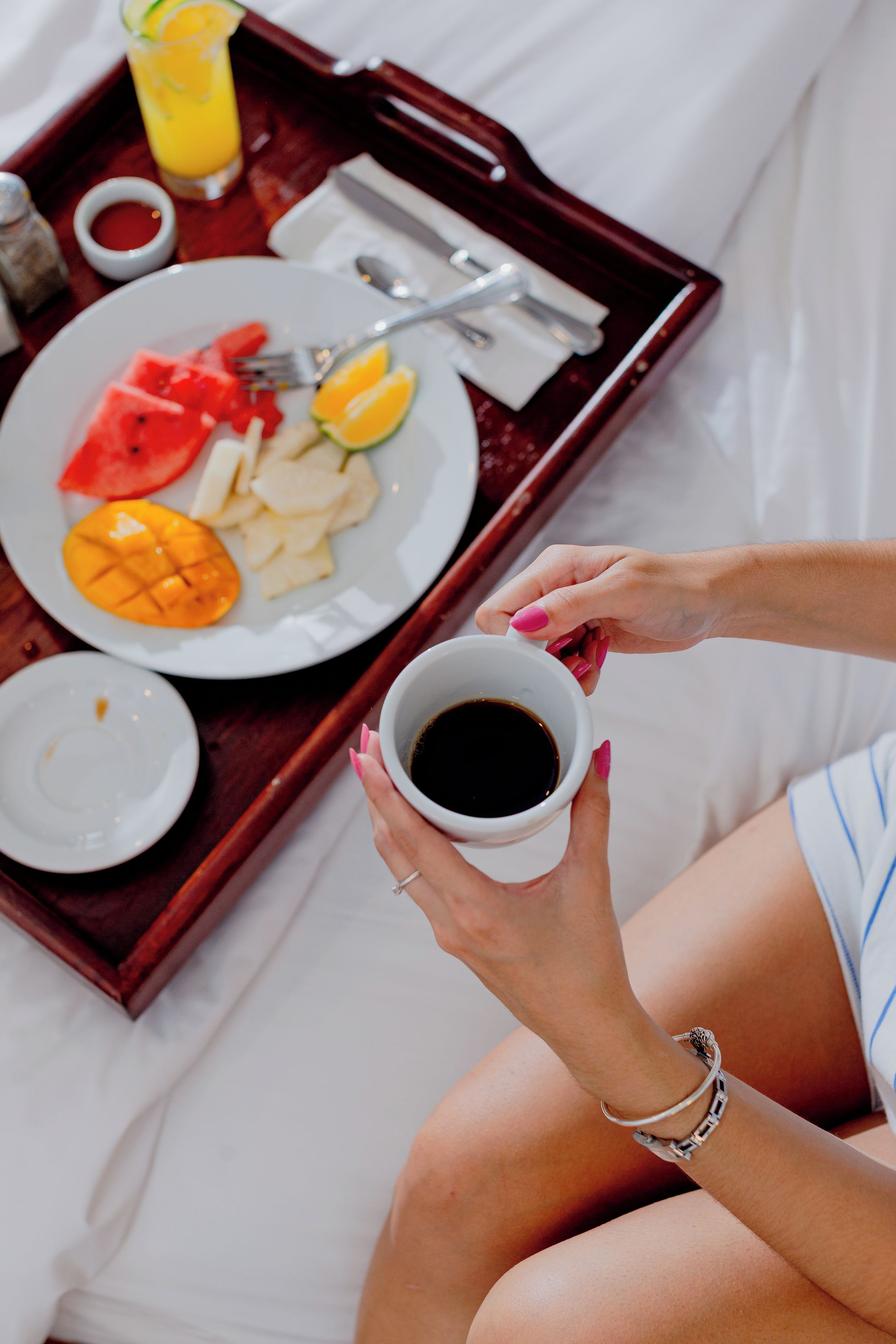 Una persona sentada en una cama con una bandeja que contiene un plato de comida, una taza de café y un vaso de jugo de naranja.