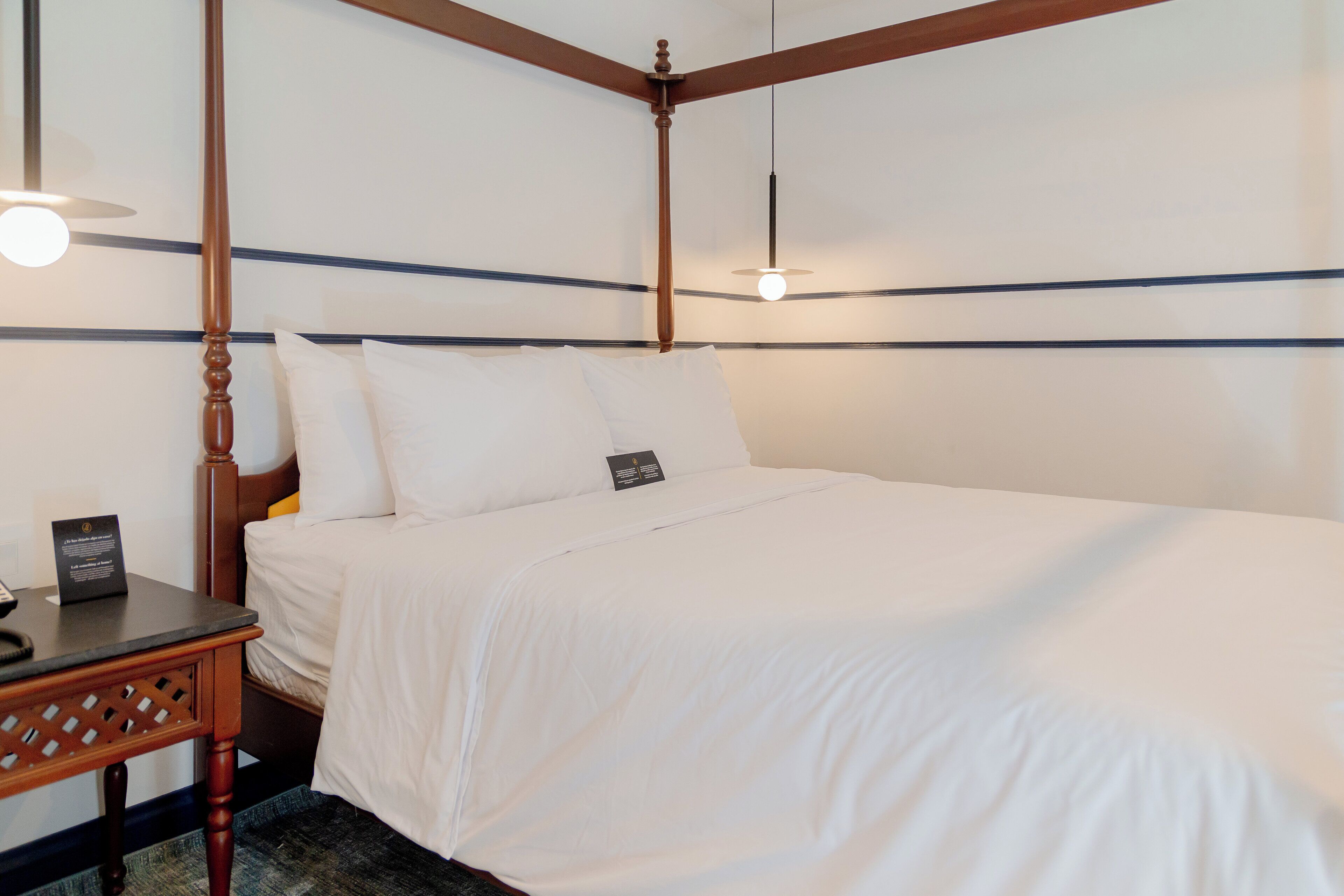 A four-poster bed with white linens, a wooden nightstand with a phone and a sign, and a hanging light fixture.