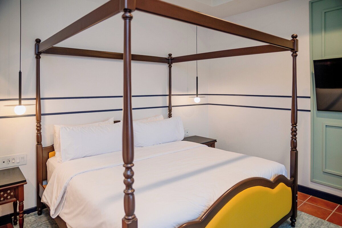 Een hemelbed met witte beddengoed, een houten hoofdbord en een nachtkastje met een lamp.