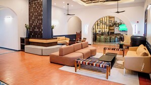 A modern lobby with a sectional sofa, a coffee table, and a seating area with chairs. There is a TV mounted on the wall and a ceiling fan.