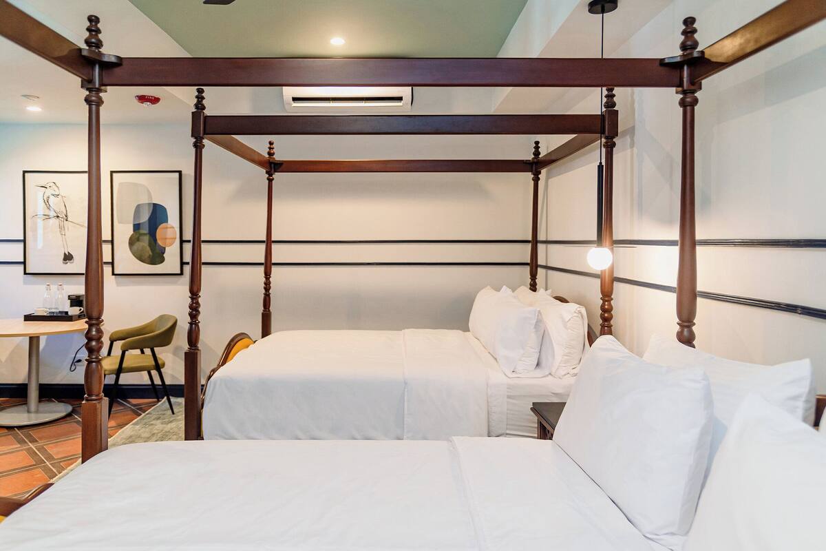 Une chambre d’hôtel avec un lit à baldaquin, deux lits simples, un bureau avec une chaise et des œuvres d’art accrochées au mur.