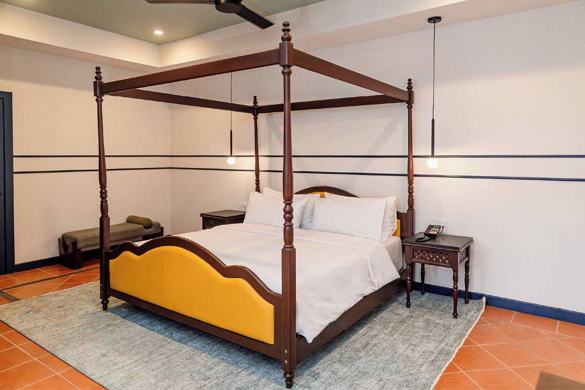 Un lit à baldaquin avec une structure en bois et une tête de lit jaune, une table de chevet avec un téléphone, et un banc adossé au mur.