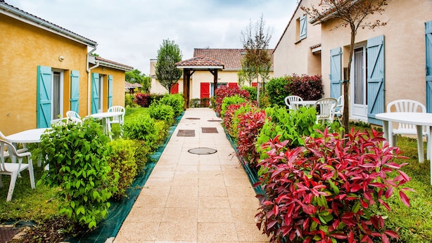 A narrow paved pathway flanked by lush greenery and white outdoor furniture, leading to a building with a tiled roof and blue shutters. - Résidence Saskia (Cazaubon)