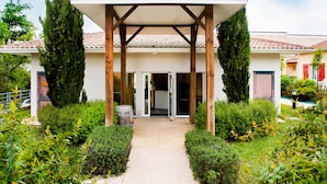 A covered outdoor area with a tiled roof, surrounded by greenery and a pathway leading to a building entrance. - Résidence Saskia (Cazaubon)