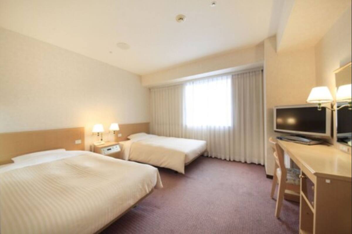 Habitación de hotel con dos camas, un escritorio con computadora, televisor y ventana con cortinas.