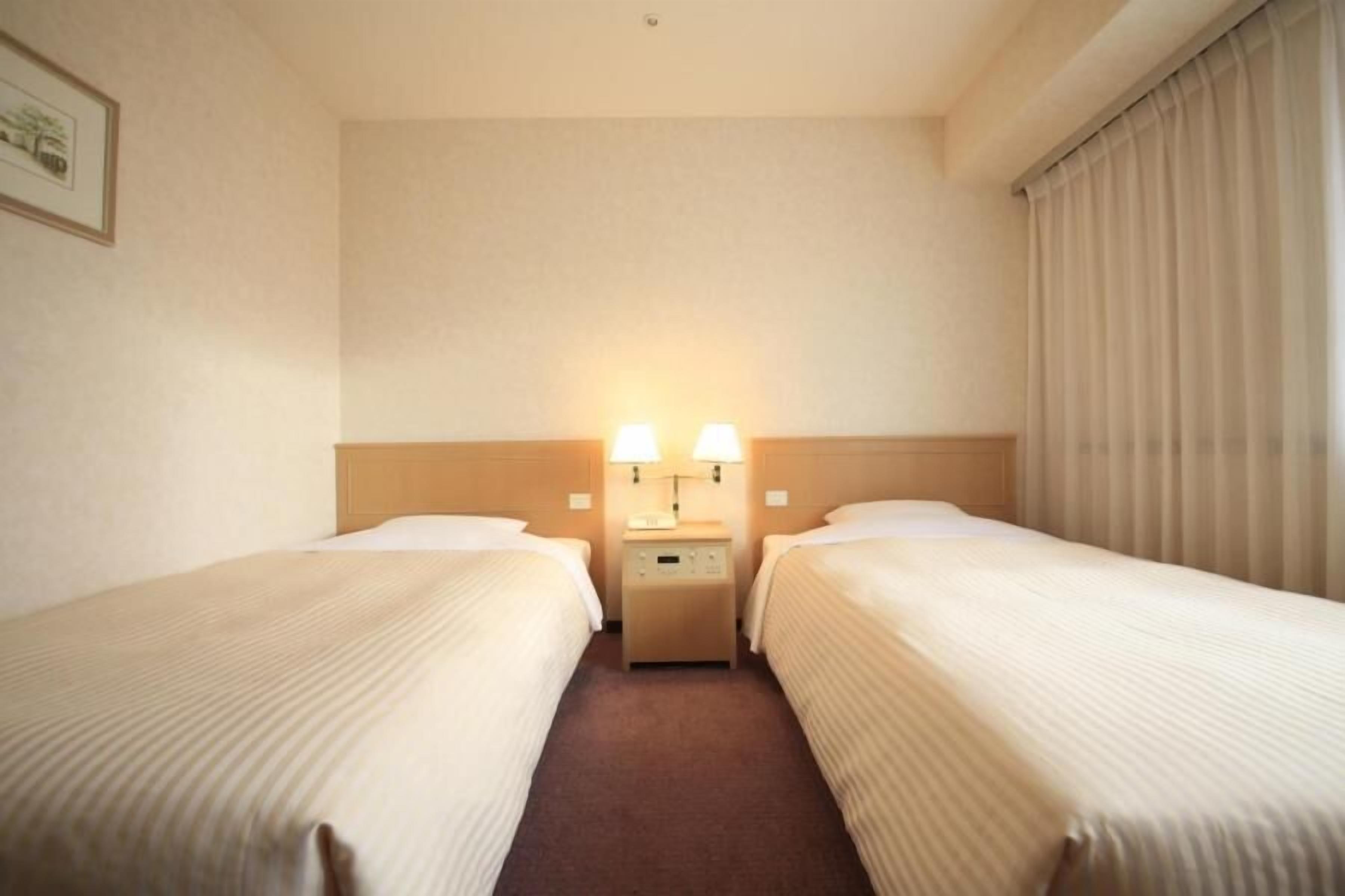 Une chambre d’hôtel avec deux lits simples, une table de chevet avec une lampe et une fenêtre avec des rideaux.