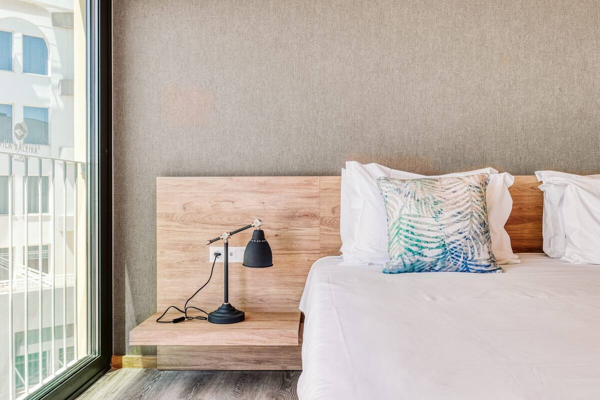 Une chambre d’hôtel avec une tête de lit en bois, une table de chevet avec une lampe, un lit avec une literie blanche et une vue sur un bâtiment à l’extérieur.