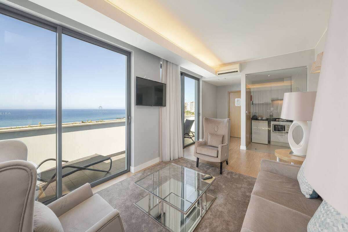 Une chambre d’hôtel moderne dotée d’un balcon, d’une télévision à écran plat, d’un canapé, d’une table basse en verre et d’un coin cuisine.