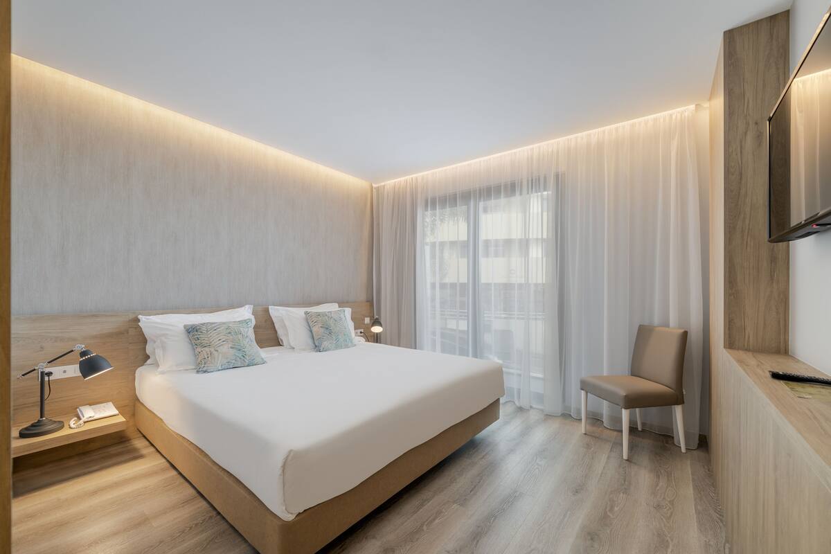 Une chambre d’hôtel moderne avec un grand lit, des tables de chevet, une chaise et une grande fenêtre avec des rideaux.